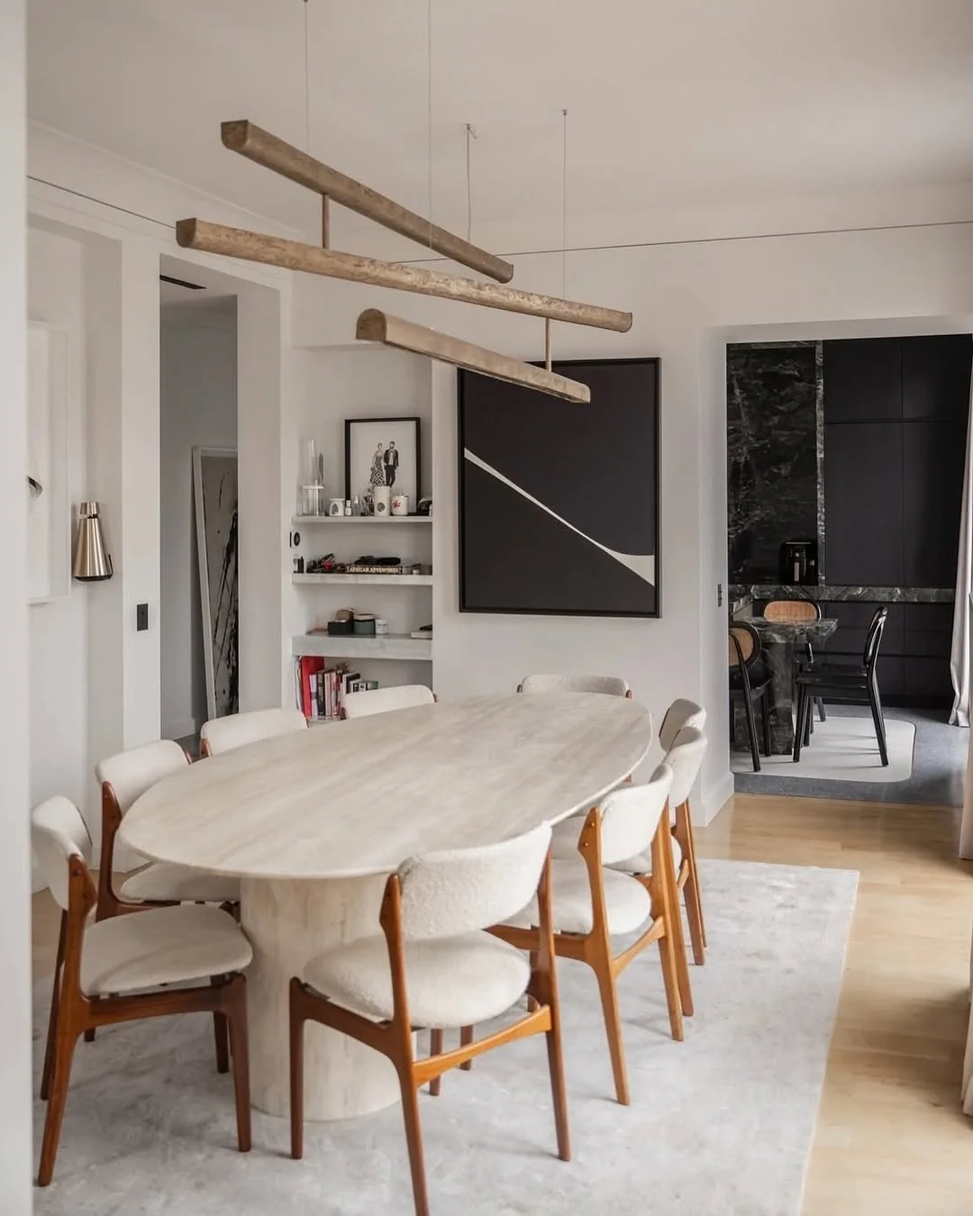 Salle à manger avec un grand table ovale en pierre et huit chaises en bois et tissu blanc, décoration moderne avec peinture abstraite noire et blanche, et suspension en bois au plafond, cuisine en arrière-plan