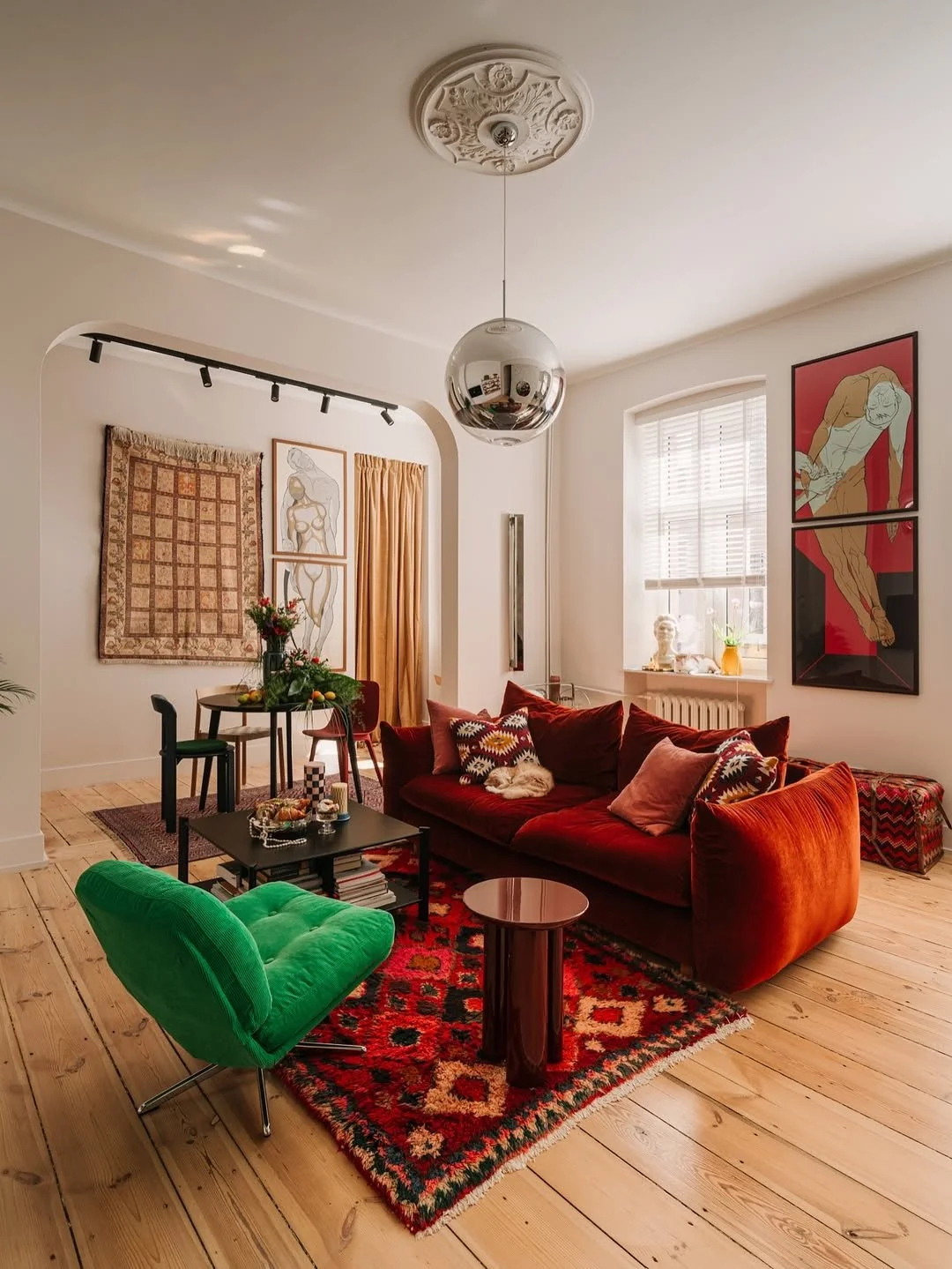 Salon vivant avec canapé rouge, fauteuil vert, tapis coloré, œuvres d'art modernes aux murs, meubles décoratifs, et une lumière naturelle passant par une fenêtre.