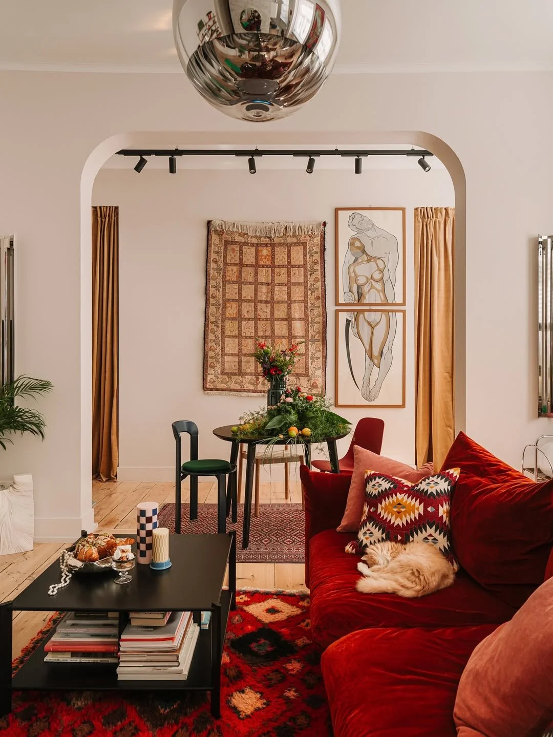 Un salon avec un canapé rouge, un tapis coloré, une table basse avec des livres, et une décoration murale dans une pièce chaleureuse.