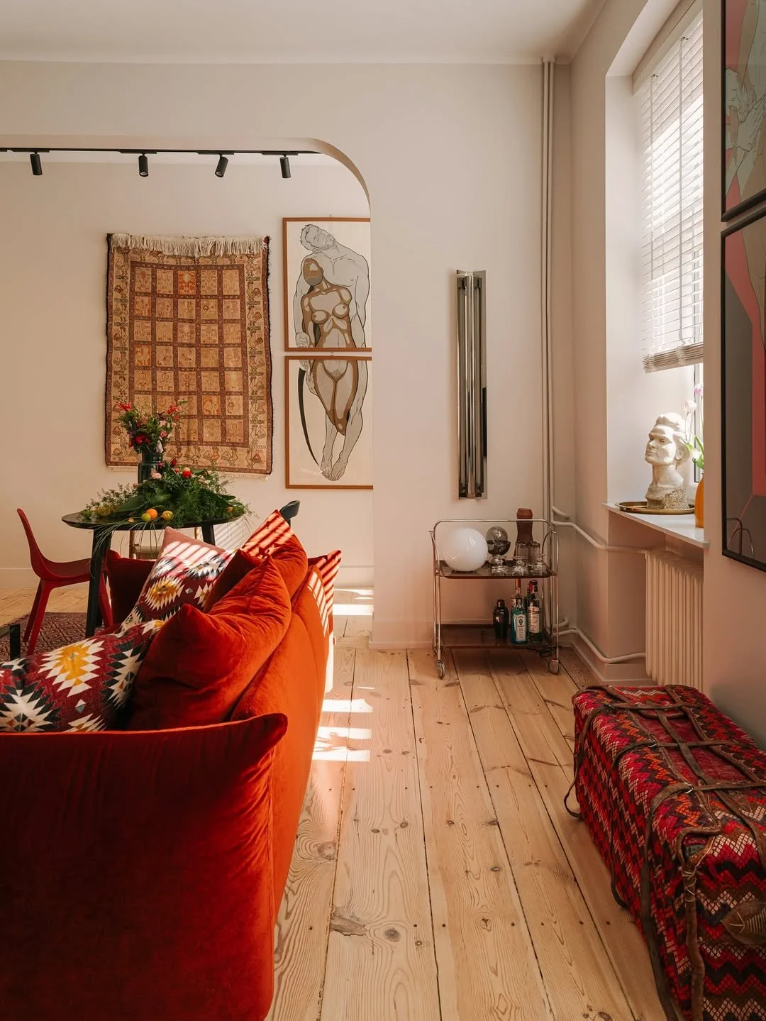 Salon décoré avec un canapé rouge, coussins colorés, un tapis, une table avec des fleurs, des tableaux modernes, une statue sur la fenêtre, une étagère avec des bouteilles, une cheminée et un coffre en tissu