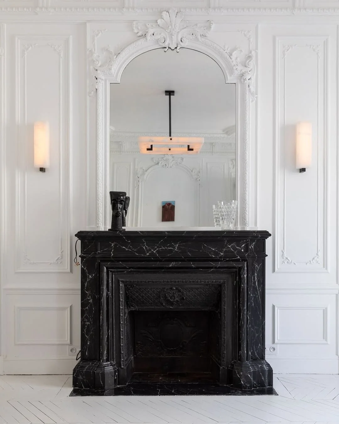 Miroir au-dessus d'une cheminée en marbre noir dans une pièce blanche élégante, avec un objet sculptural noir et un verre en cristal sur la cheminée. Deux appliques murales en bleu clair encadrent le miroir.