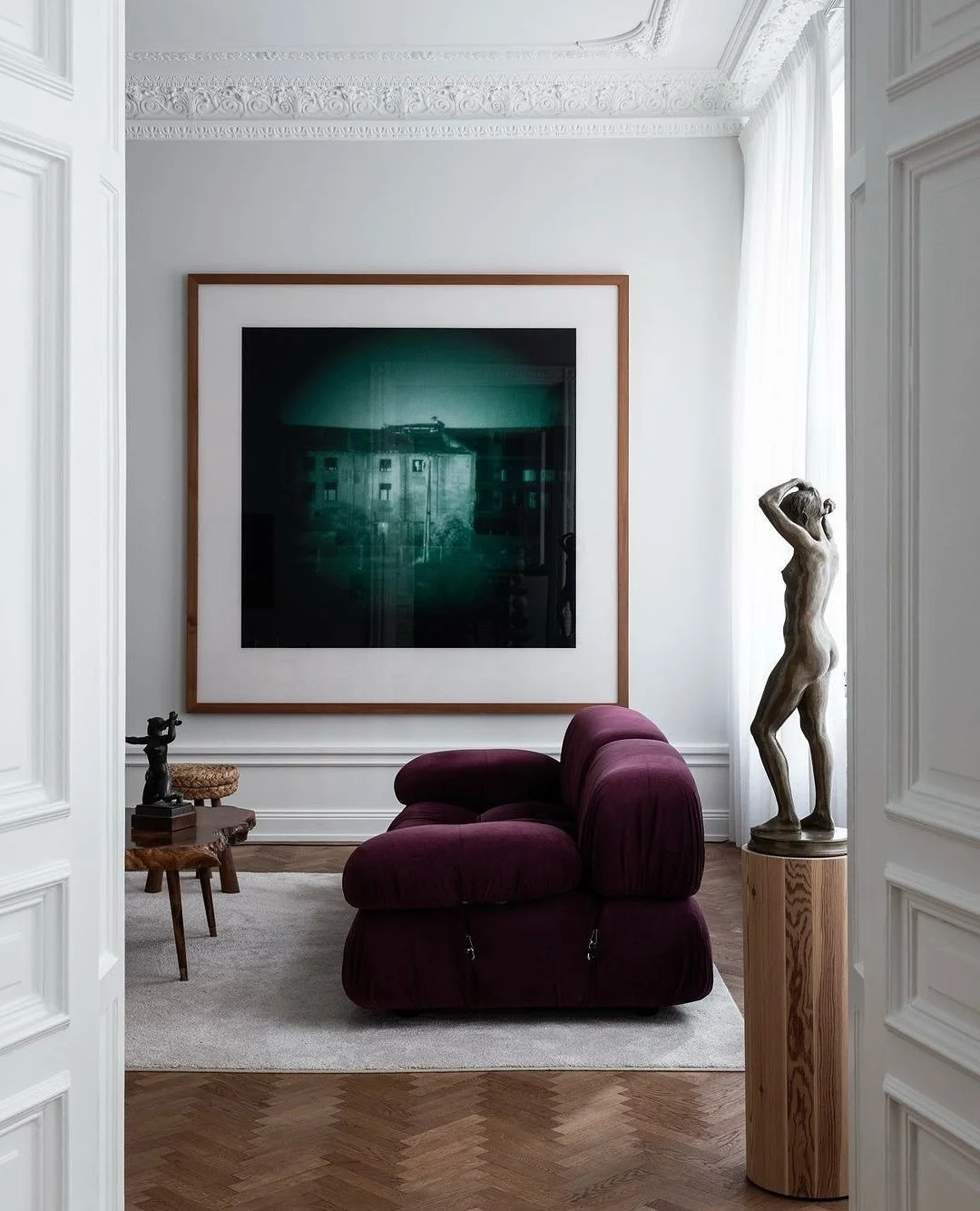 Salon élégant avec un canapé violet, une sculpture en bois d'une femme, un tableau sombre encadré en bois, une petite table en bois avec une figurine, et un rideau blanc à côté d'une fenêtre.