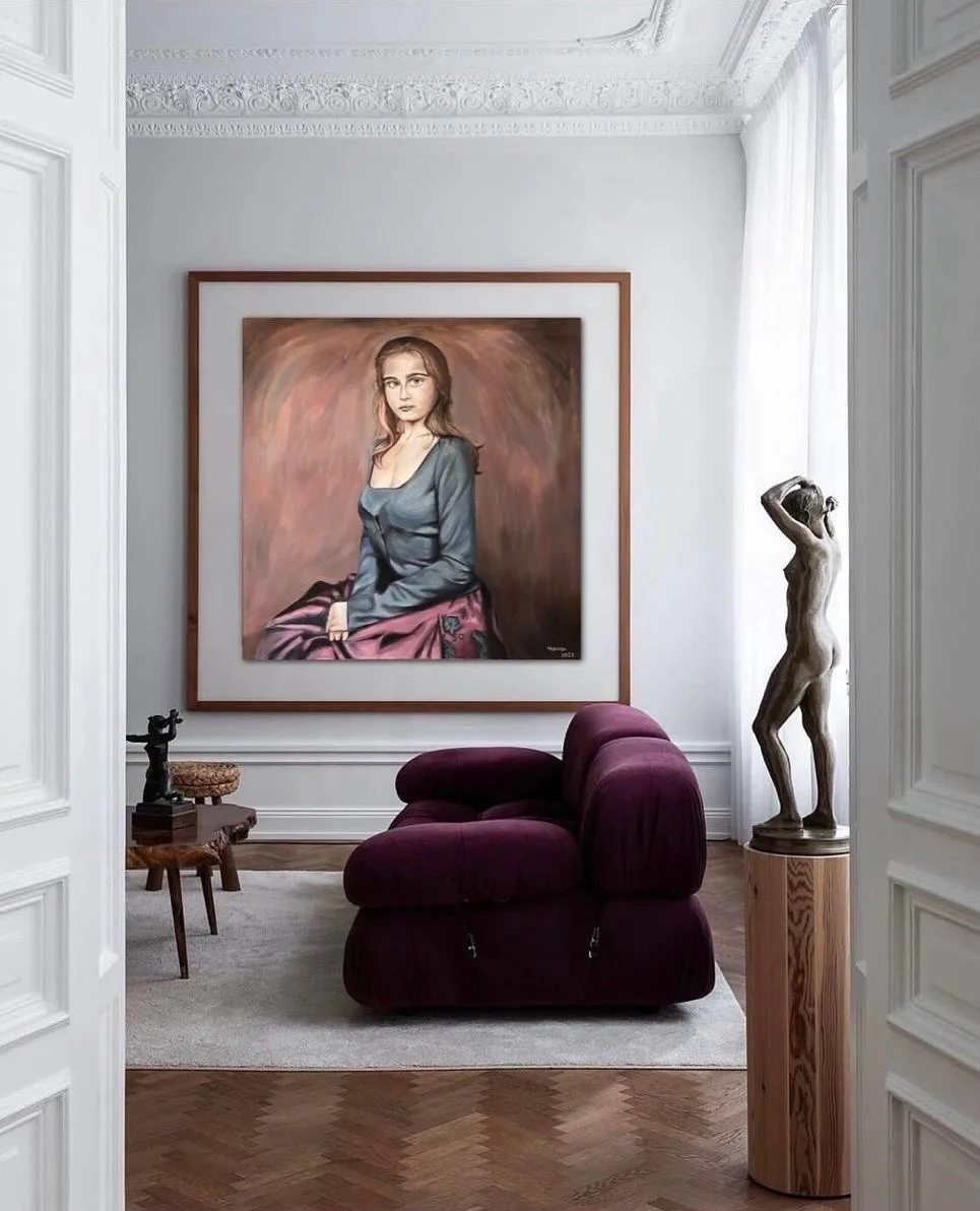 Une salle d'exposition élégante avec un tableau représentant une jeune femme, une sculpture en bois d'une femme, un fauteuil couleur bourgogne et une petite table en bois avec une sculpture. La pièce a des murs blancs, un plafond décoré et un sol en parquet.