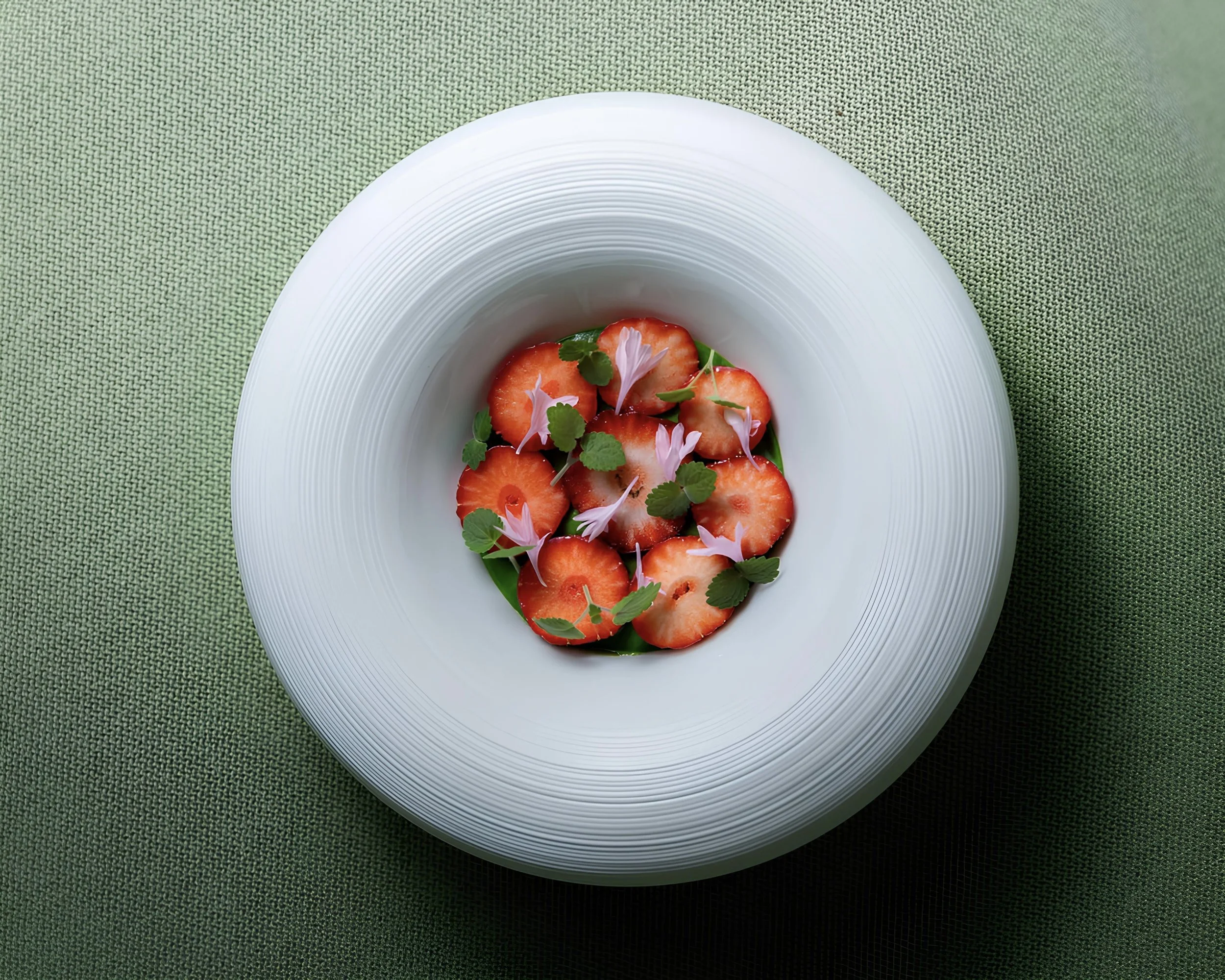 Assiette avec des tranches de fraises décorées de feuilles de menthe et de petites fleurs roses, sur une nappe verte.