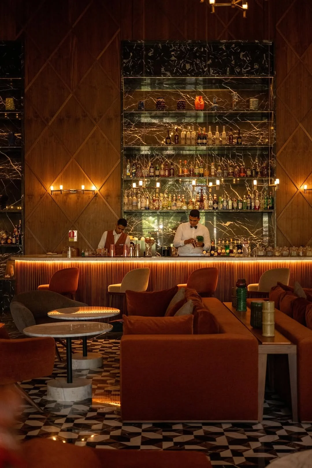 Un bar élégant avec deux barmans derrière le comptoir. L'arrière du bar est en marbre noir avec des étagères remplies de bouteilles d'alcool. La pièce est décorée avec des murs en bois et des tables et chaises confortables.