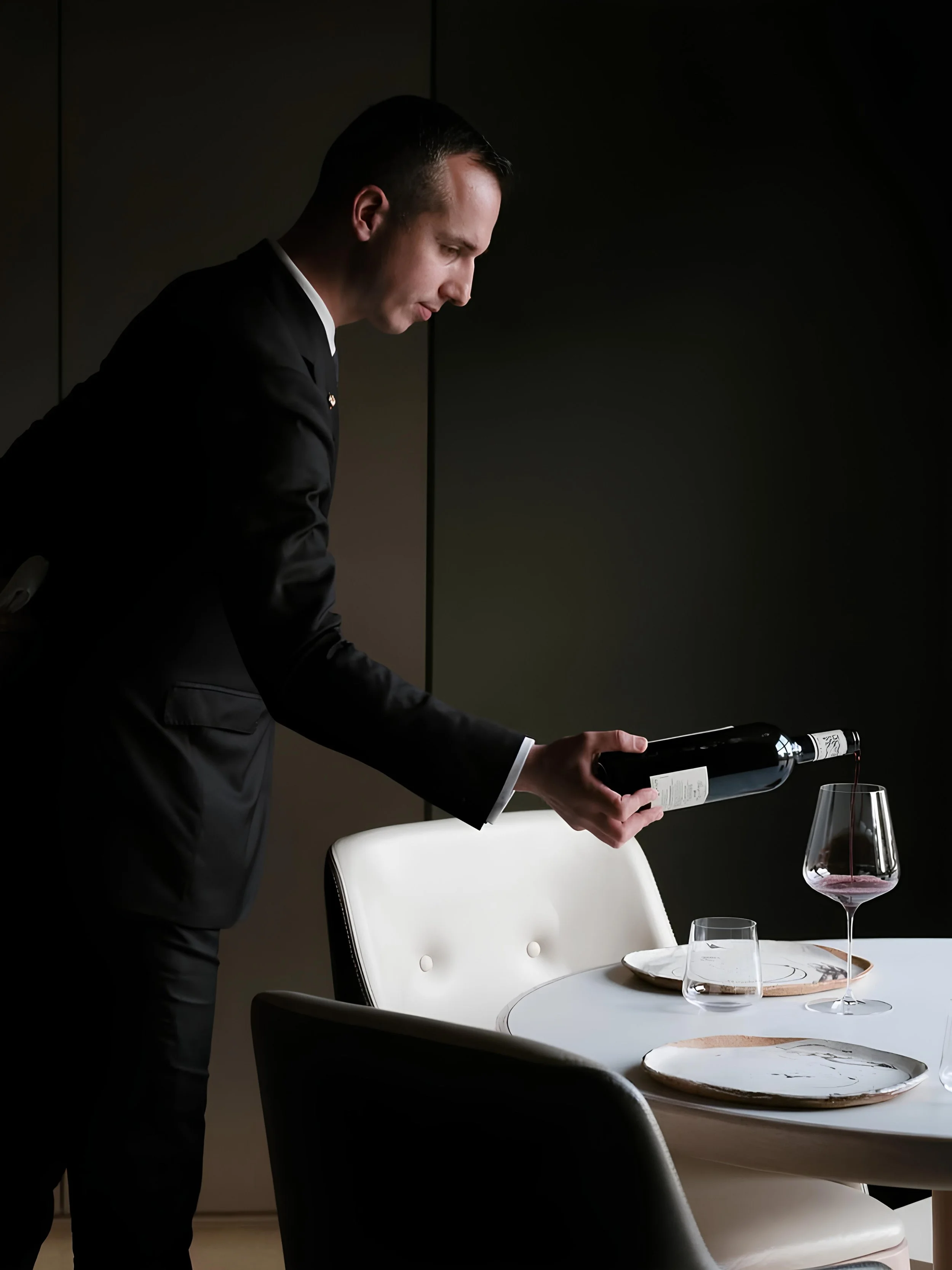 Homme en costume noir versant du vin rouge dans un verre à vin placé sur une table élégante.