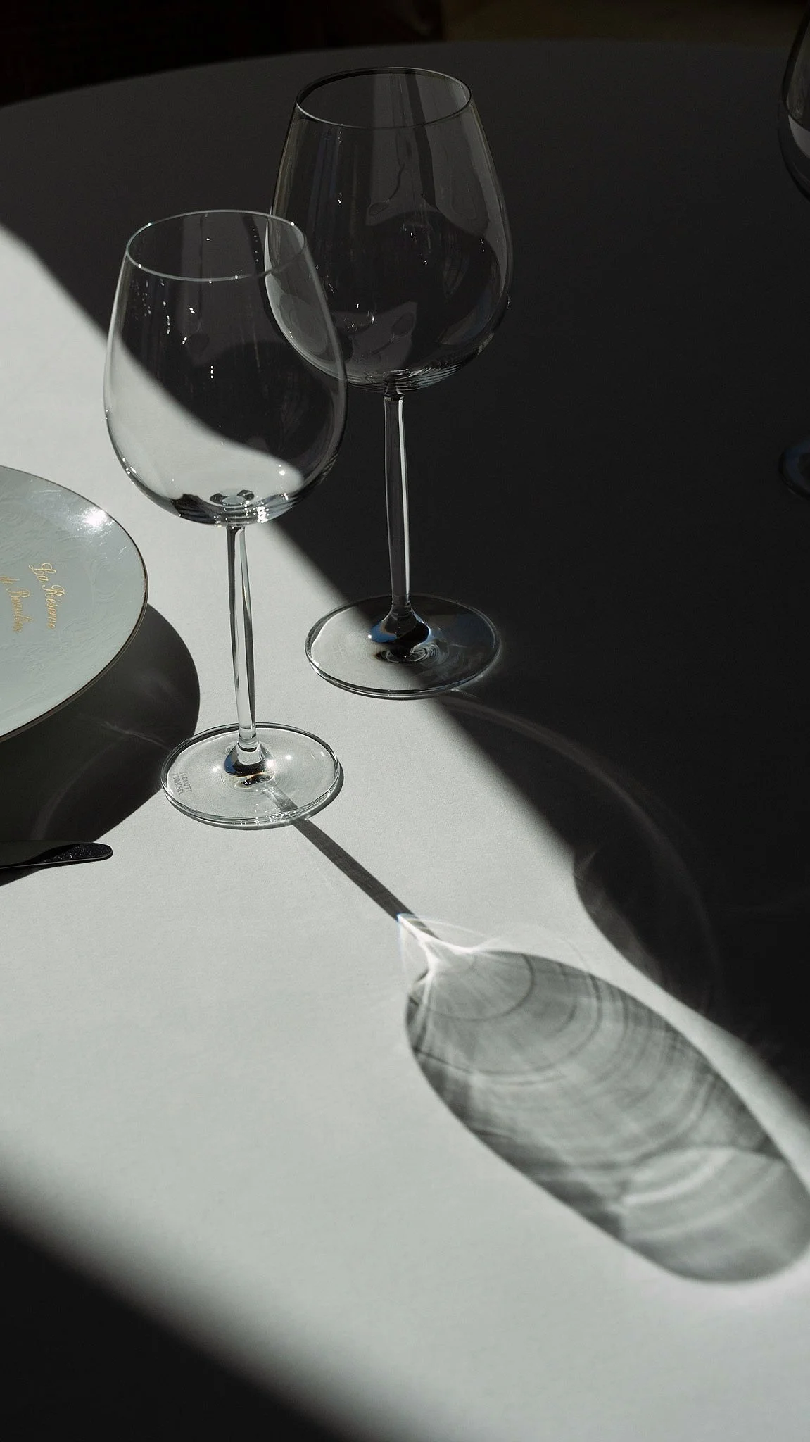 Deux verres à vin vides avec des ombres et reflets prononcés sur une surface blanche, avec un fond sombre.