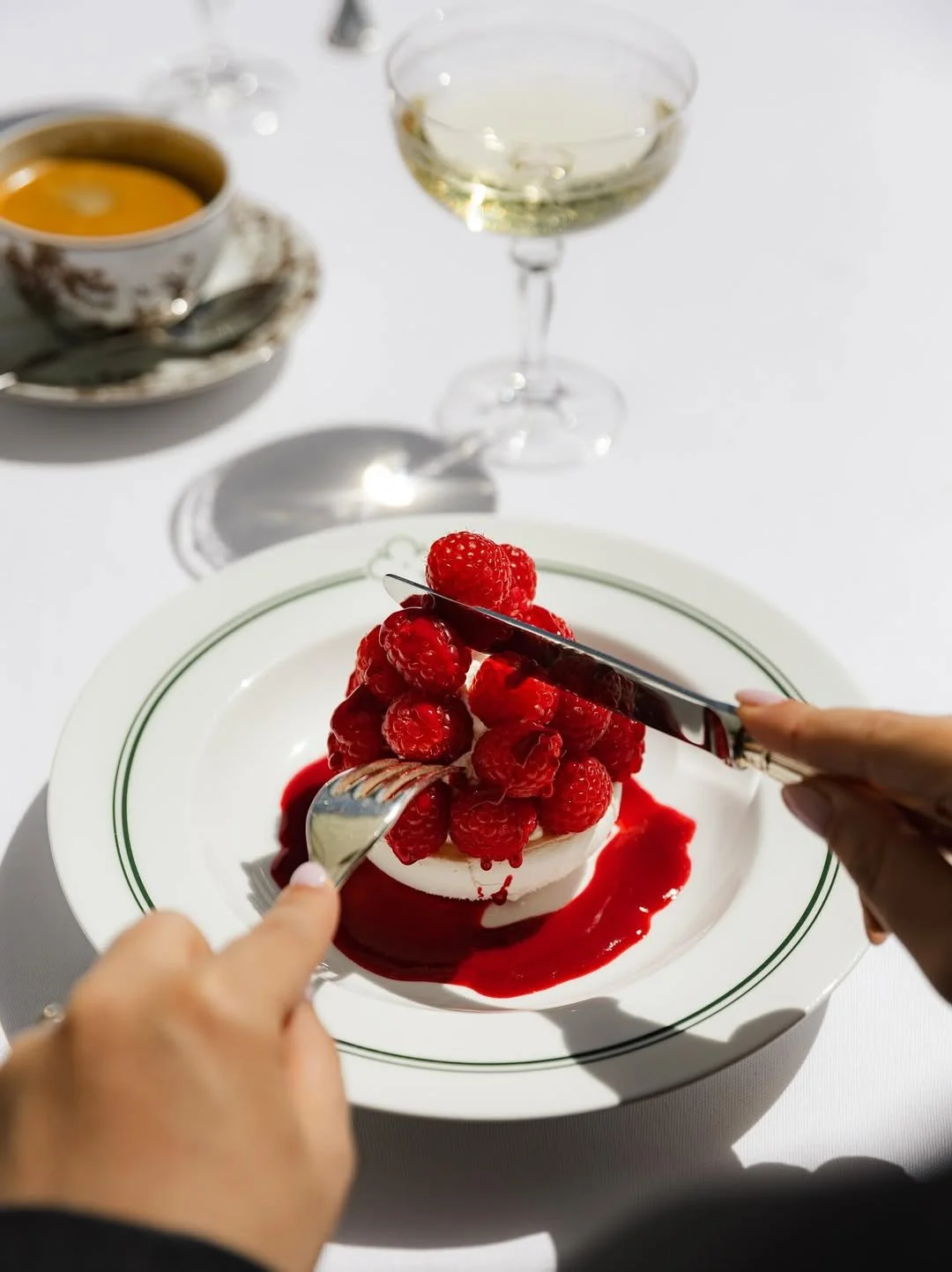 Gâteau aux framboises sur une assiette blanche, avec des couverts, une coupe de vin blanc, une tasse de café, et un verre de vin blanc en arrière-plan.