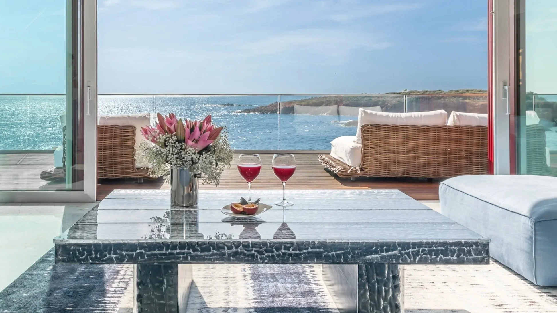 Salon avec vue sur la mer, comprenant une table en marbre avec deux verres de vin rouge, une corbeille de fleurs et un plateau de figues, avec canapés en rotin et coussins blancs sur le balcon extérieur.