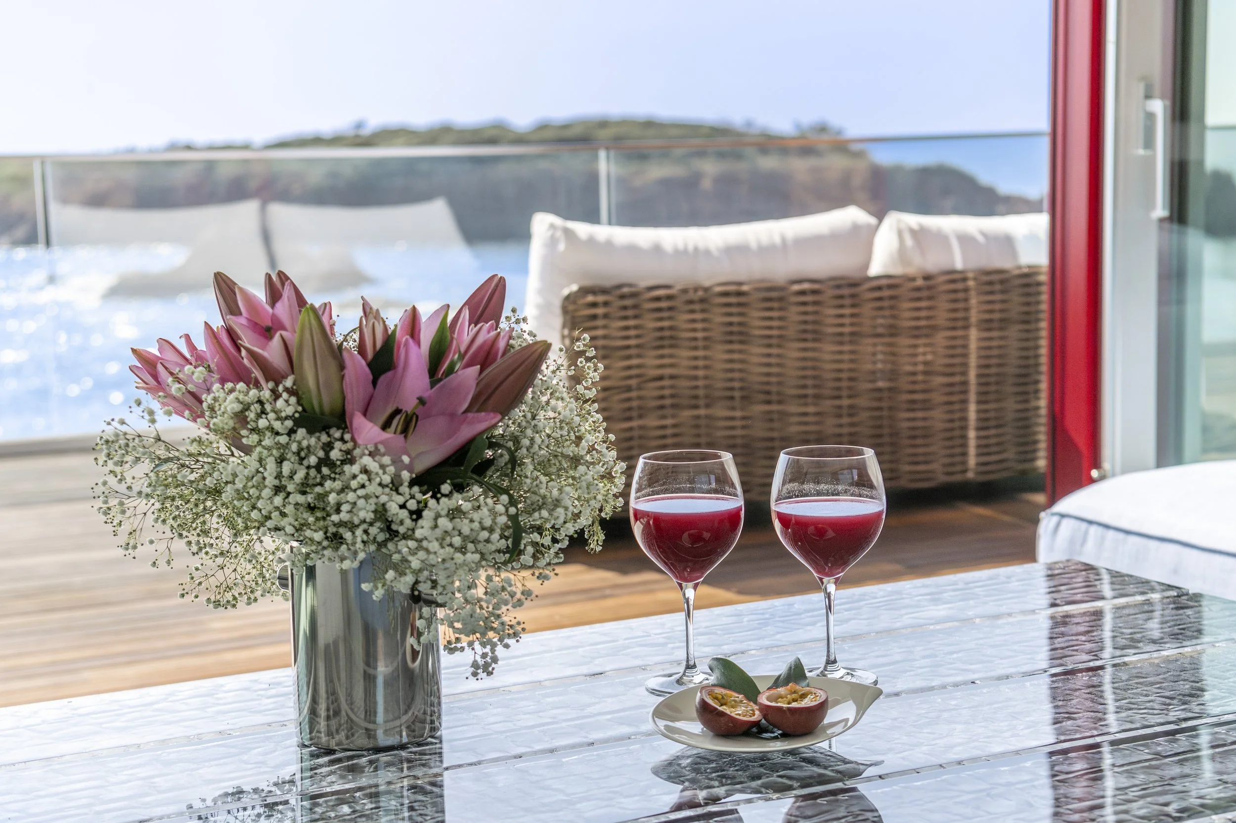 Un vase de fleurs avec des lys roses sur une table en verre, deux verres de vin rouge, et un demi-fruit sur une assiette, devant une baie vitrée avec vue sur la mer et un canapé en rotin.