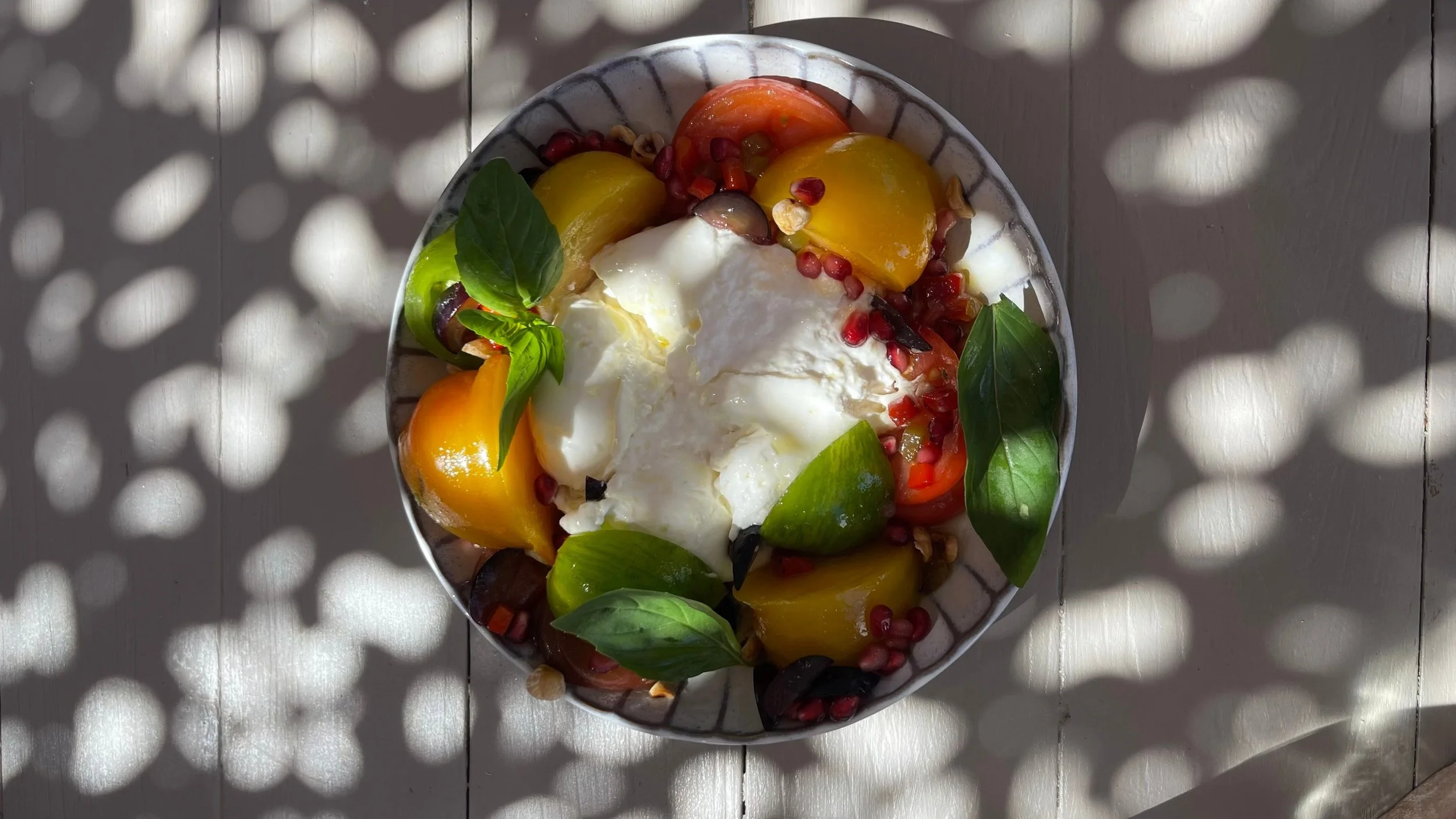 Salade caprese avec des morceaux de tomates jaunes, vertes, rouges, de la mozzarella, du basilic, des grenades et des noix, servie dans un bol blanc, avec un jeu d'ombres découpées sur la table en bois.