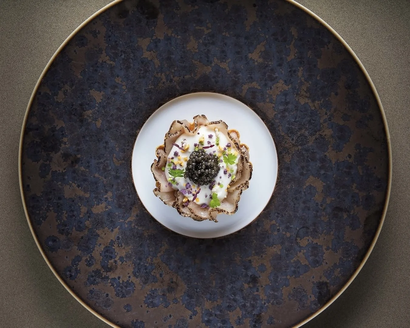 Un plat sophistiqué avec des tranches de viande autour d'une sauce blanche, garnie de caviar noir, de jeunes pousses et de fleurs comestibles, présenté sur une assiette blanche au centre d'une assiette noire.