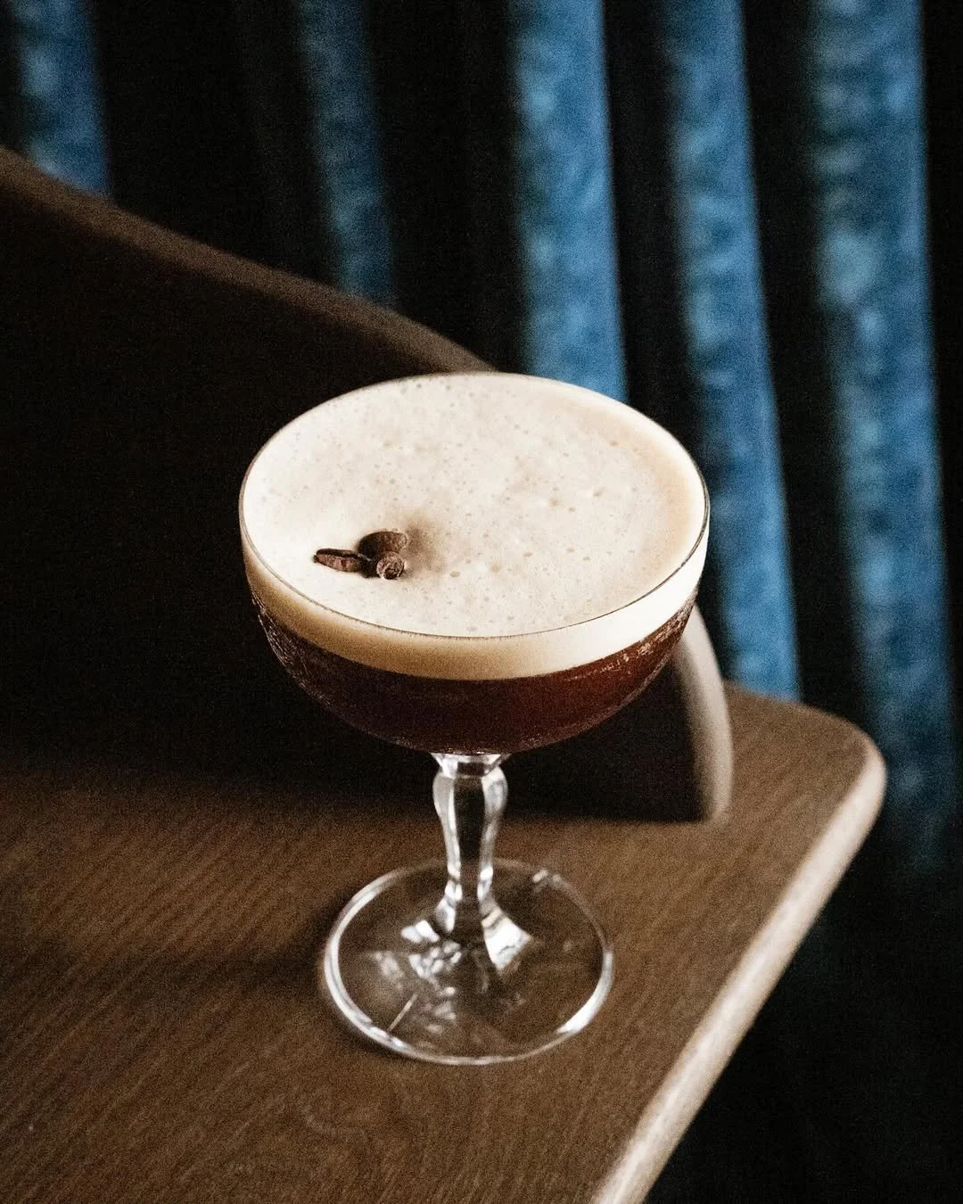 Cocktail avec une couche de mousse, décoré d'un grain de café, sur une table en bois, fond de rideaux bleus.