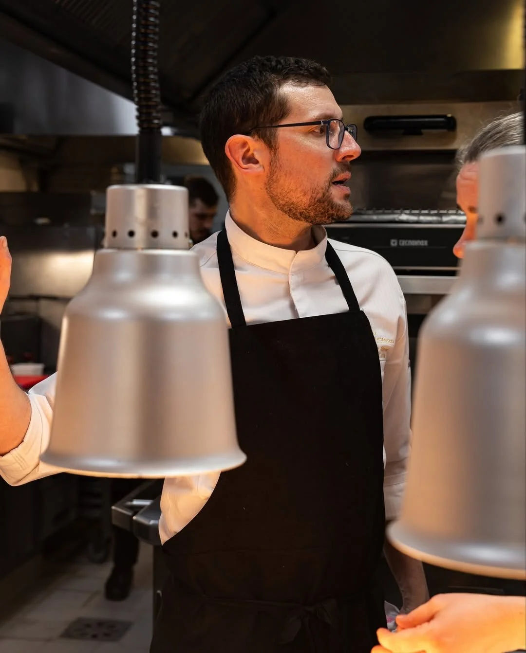 Chef en cuisine avec lunettes, portant un uniforme blanc et un tablier noir, conversant avec une personne dans une cuisine professionnelle.
