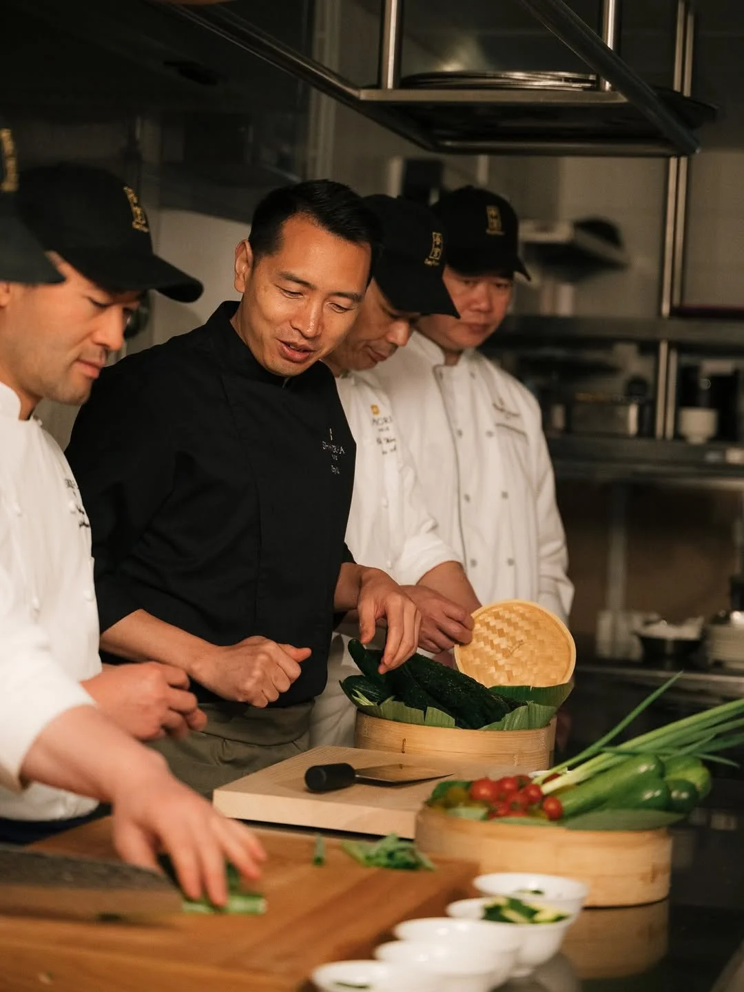 Un chef asiatique avec plusieurs assistants en uniforme blanc et casquettes noires préparant des légumes dans une cuisine professionnelle, notamment des concombres et des oignons verts.