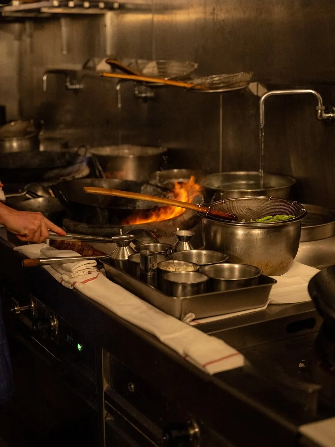 Cuisine de restaurant avec une cuisinière en train de faire sauter des aliments avec un feu vif, plusieurs casseroles en acier inoxydable et des ustensiles de cuisine.