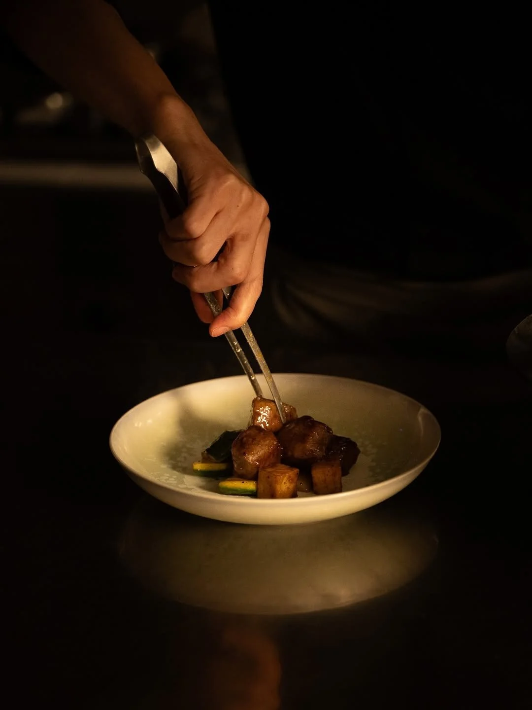 Un chef garnissant un plat avec des morceaux de viande et légumes, utilisant des pinces dans une cuisine sombre.