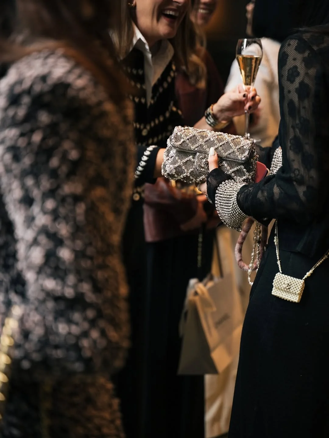 Groupe de personnes lors d'une fête ou d'un événement social, tenant des verres de champagne et portant des tenues élégantes, avec un focus sur une femme tenant un sac à main orné de perles.