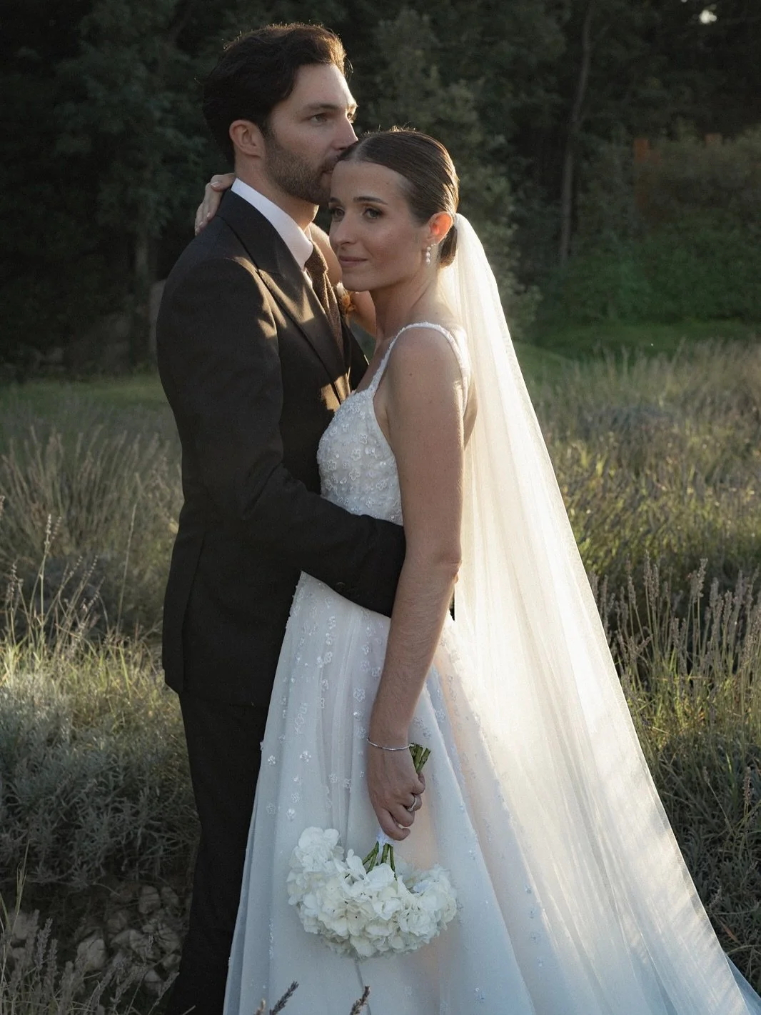 Un couple en robes de mariage et costume noir, se tenant dans un champ lors d'un mariage, avec la femme tenant un bouquet blanc.