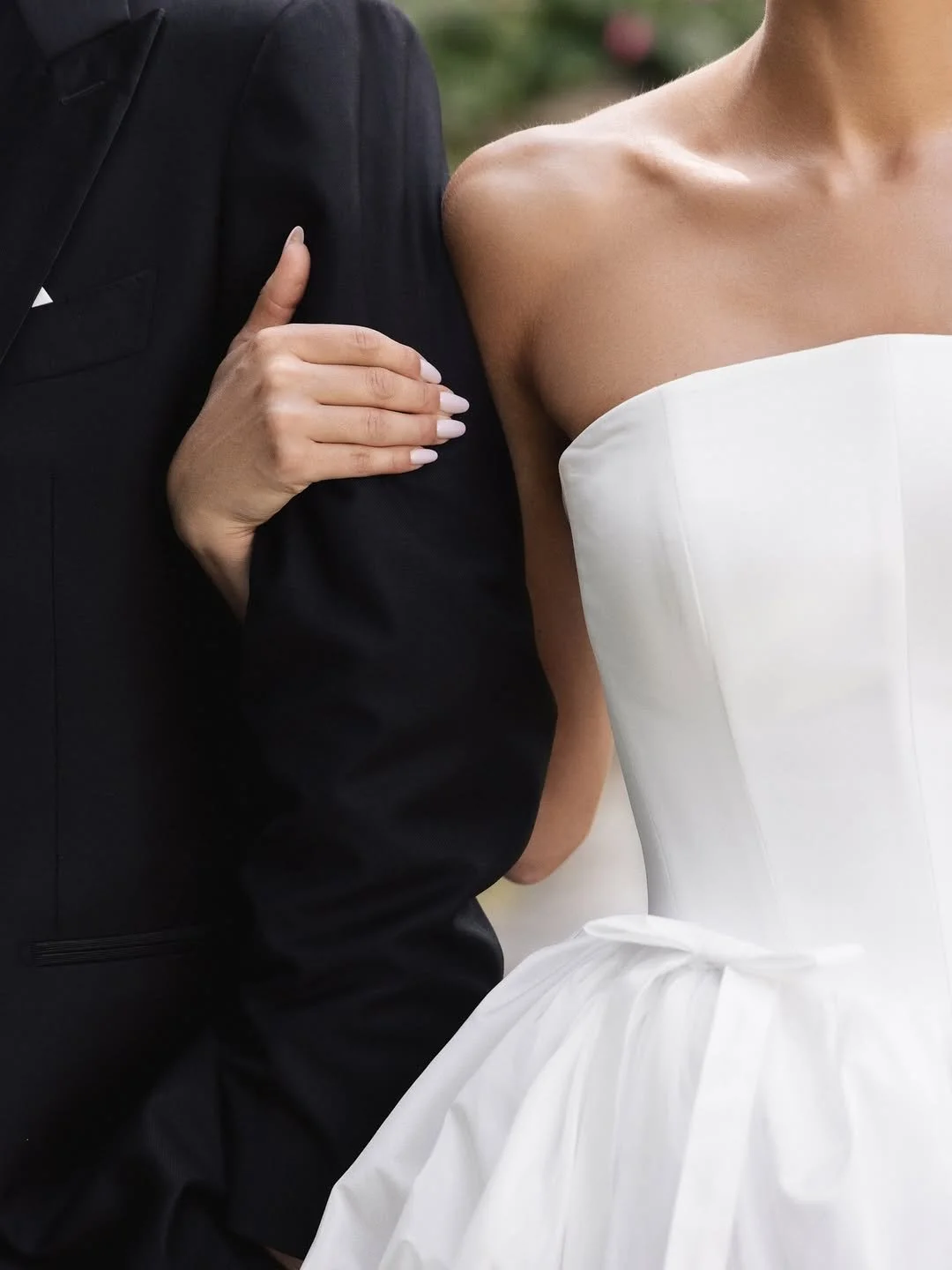 Détail d'un couple en vêtement de mariage, la femme portant une robe blanche et l'homme en costume noir, avec la main de la femme posée sur le bras de l'homme.