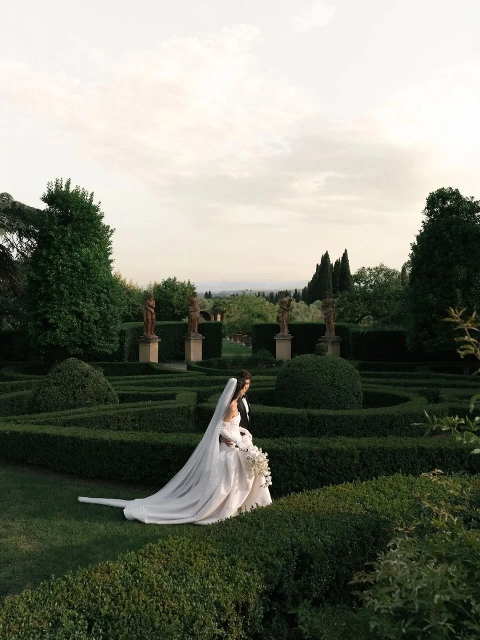 Une mariée en robe blanche avec un voile long assise dans un jardin bien entretenu avec des statues et des topiaires.
