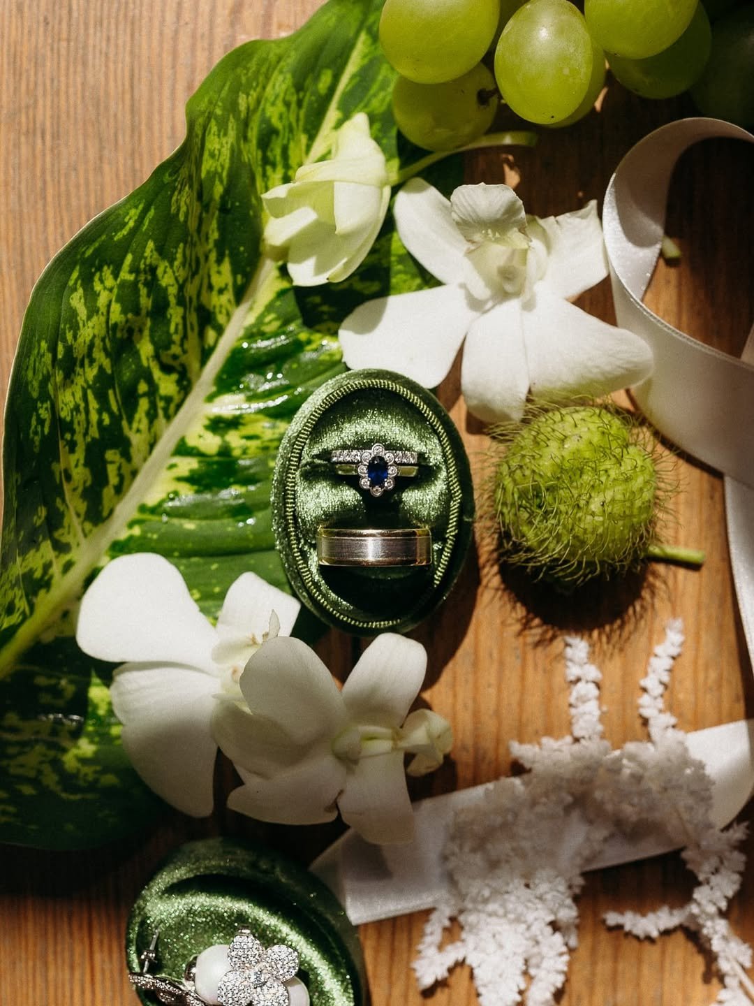 Une composition florale avec des feuilles vertes, des fleurs blanches, des grappes de raisin vert, un fruit jaune spongieux, et des bijoux (une bague avec une pierre bleue, une bague avec des diamants en forme de cœur, et une bague en argent) dans un décor en bois avec une boucle en ruban blanc.