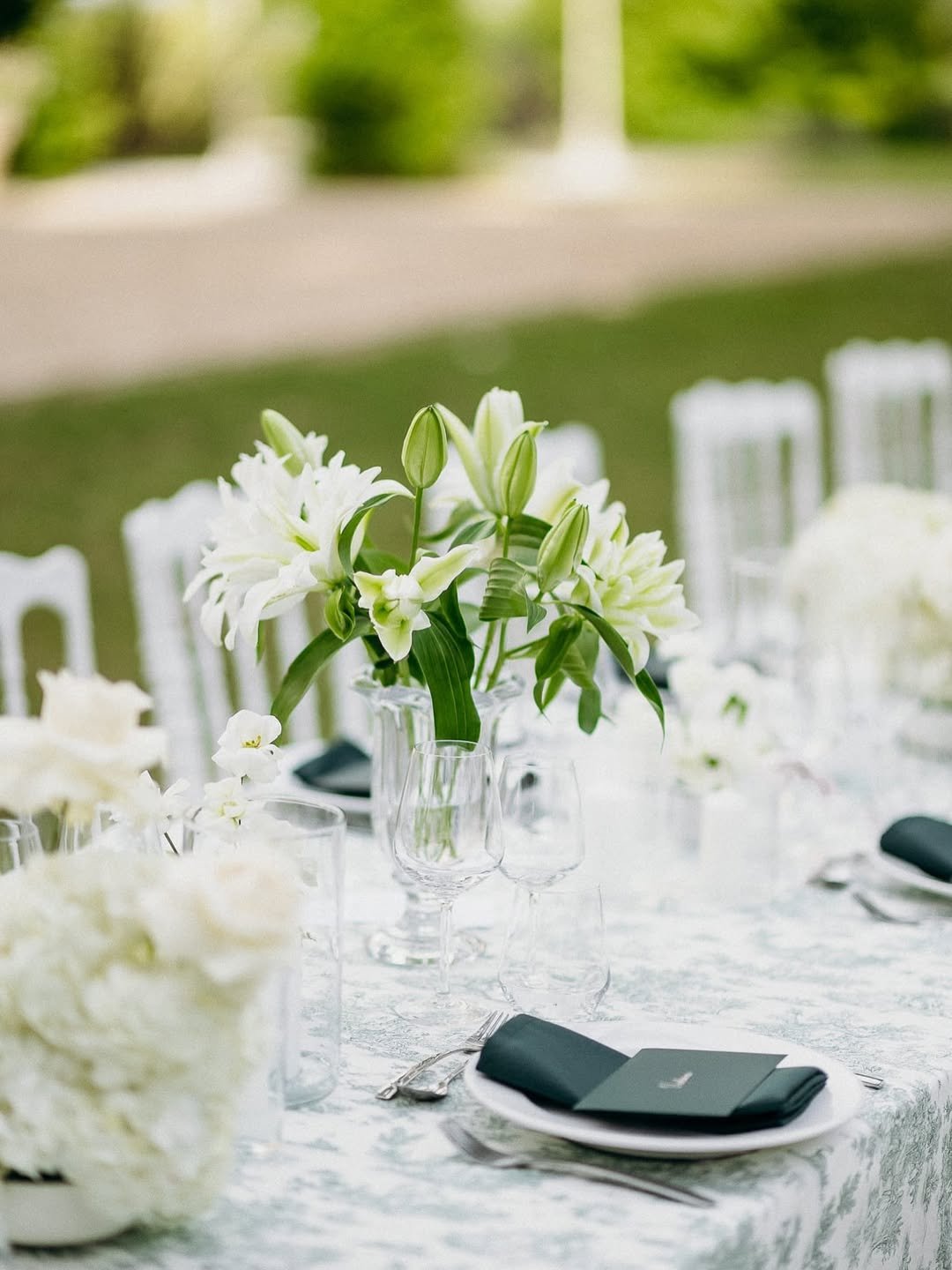 Table de mariage élégante avec une nappe blanche, un centre de table de fleurs blanches, plusieurs verres à vin, des assiettes avec des serviettes noires, et des chaises blanches en arrière-plan, en plein air avec un jardin verdoyant.