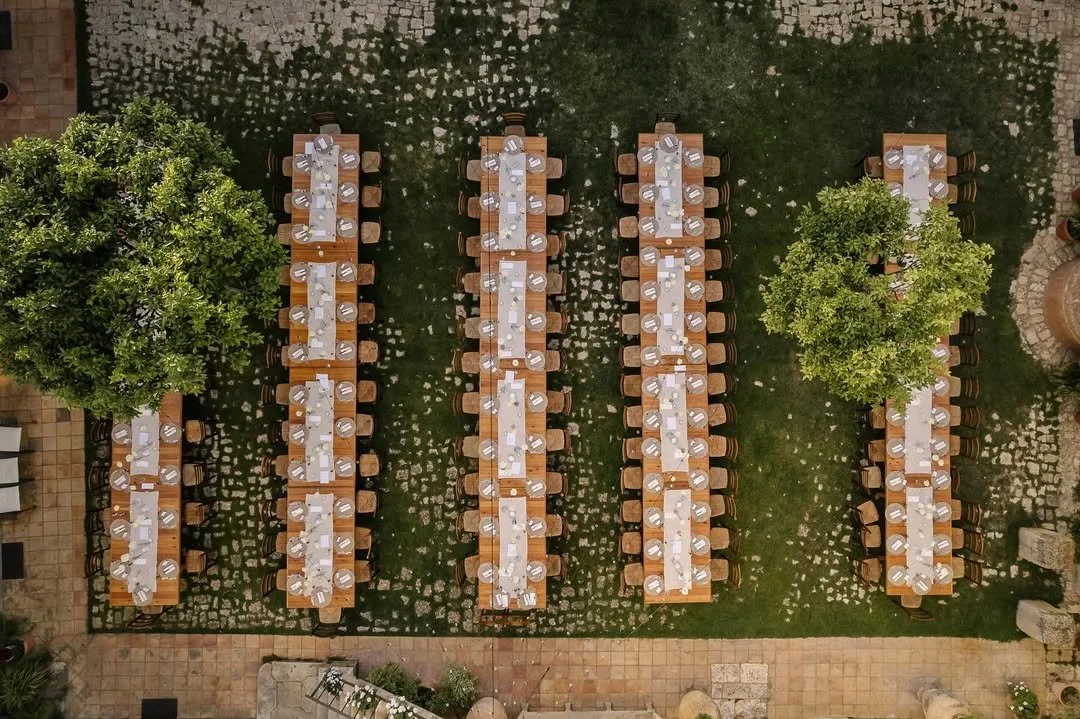 Disposition de quatre longues tables en bois avec nappe blanche, assiettes, verres et couverts, dans un espace extérieur avec deux grands arbres et un sol en pierre.