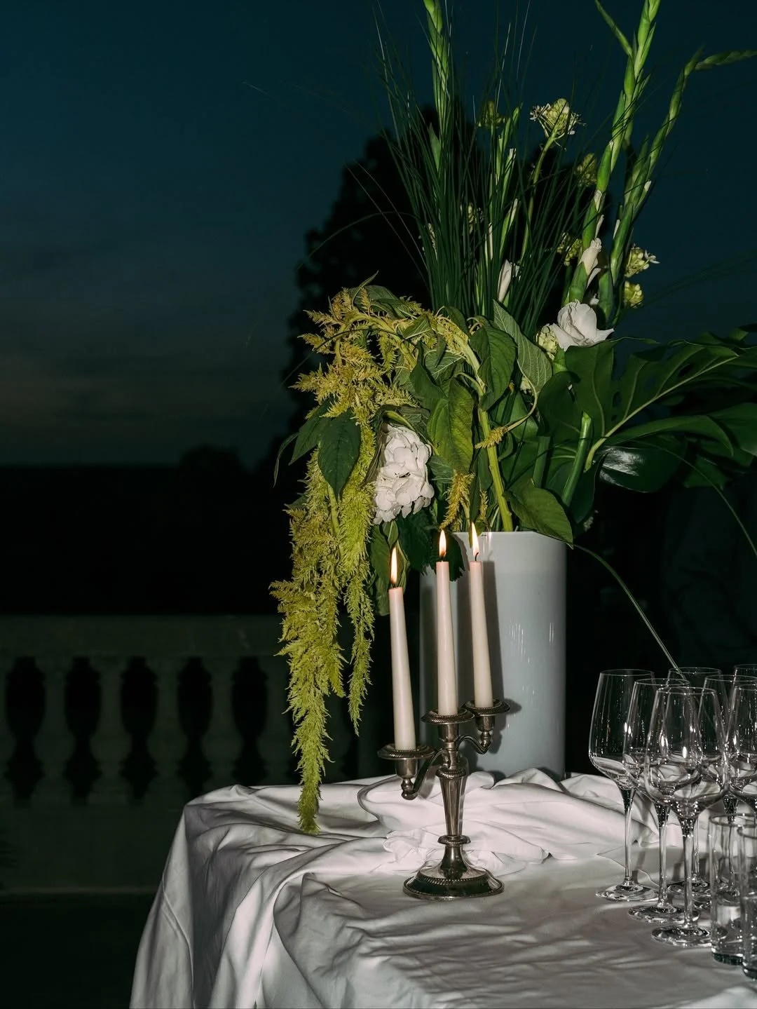 Vase avec grandes plantes vertes et fleurs blanches sur une table avec une étoffe blanche, bougie en chandelier en argent et verres à vin vidés.