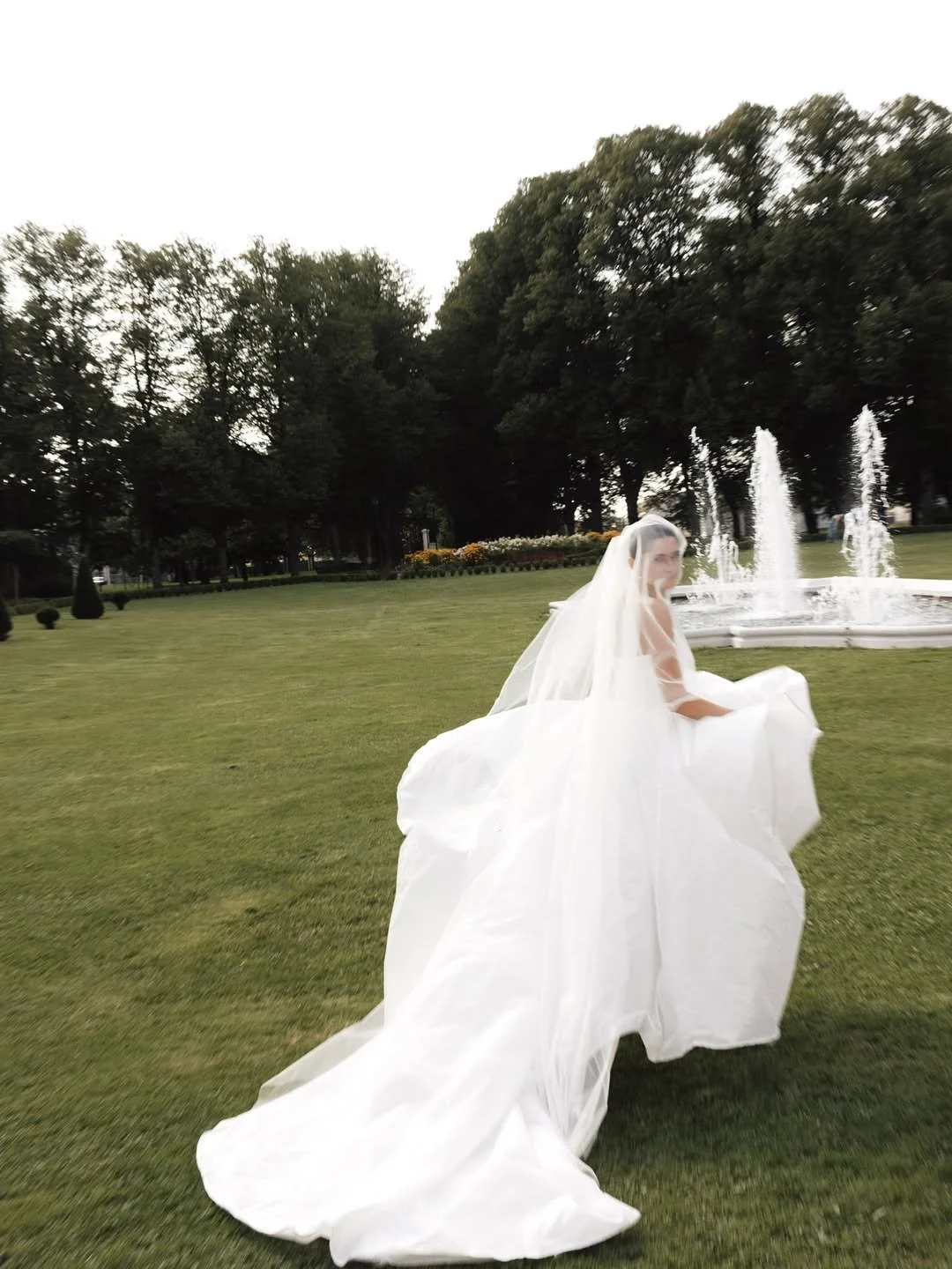 Une femme en robe de mariée avec un voile blanc se tenant dans un parc avec un jet d'eau en arrière-plan.
