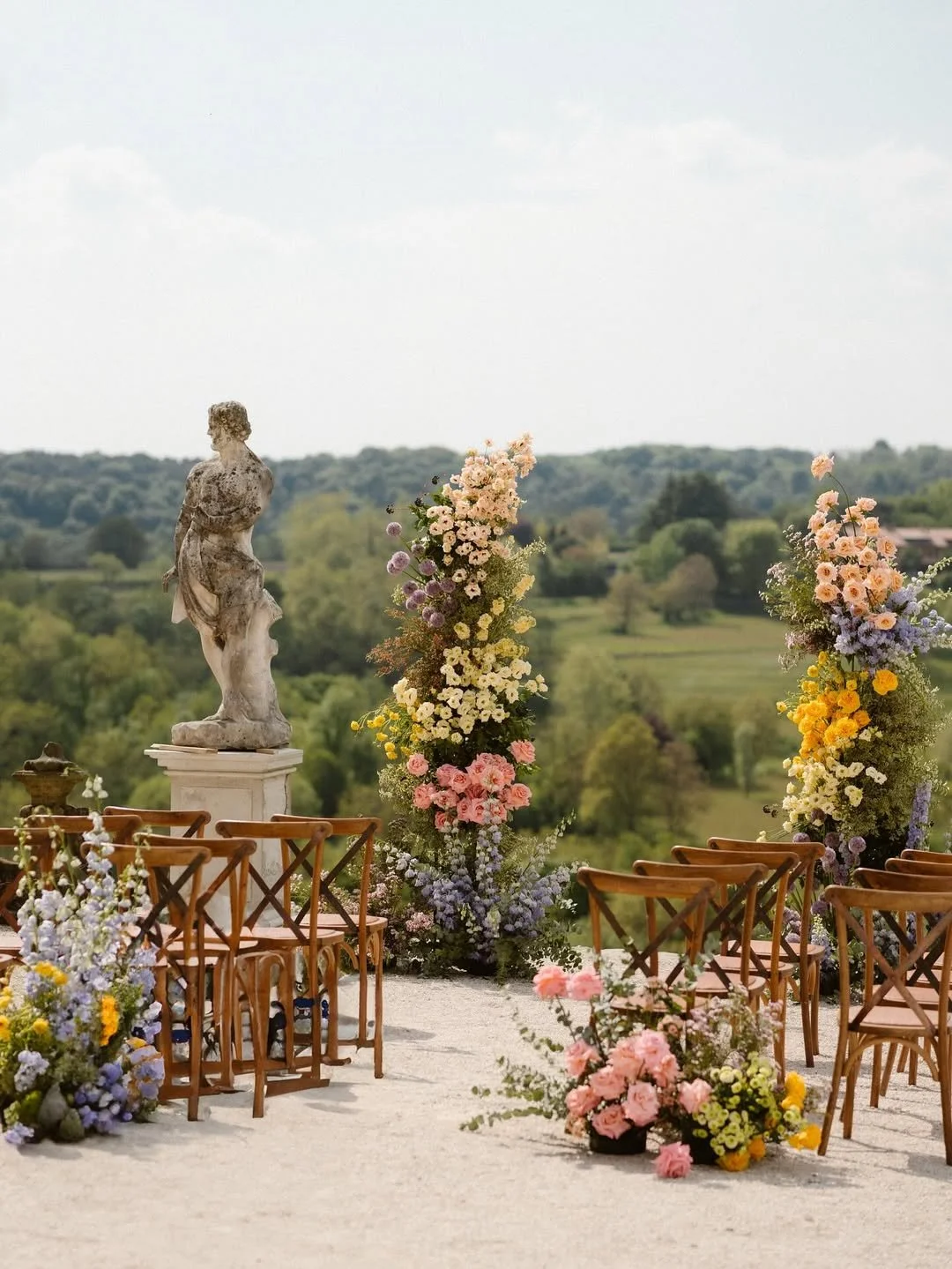 Décoration extérieure de mariage avec des fleurs pastel, une statue antique, et un paysage verdoyant en arrière-plan.