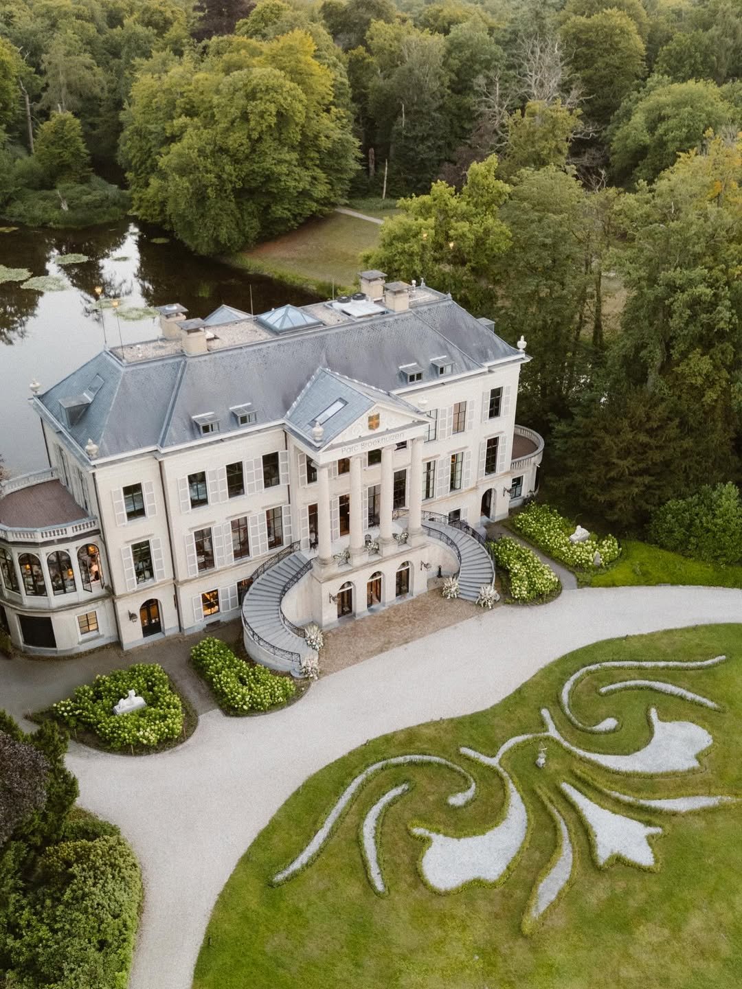 Un grand château blanc avec des colonnes, entouré d'un jardin bien entretenu avec des formes en gravier et des fleurs, situé près d'un lac et entouré d'arbres.