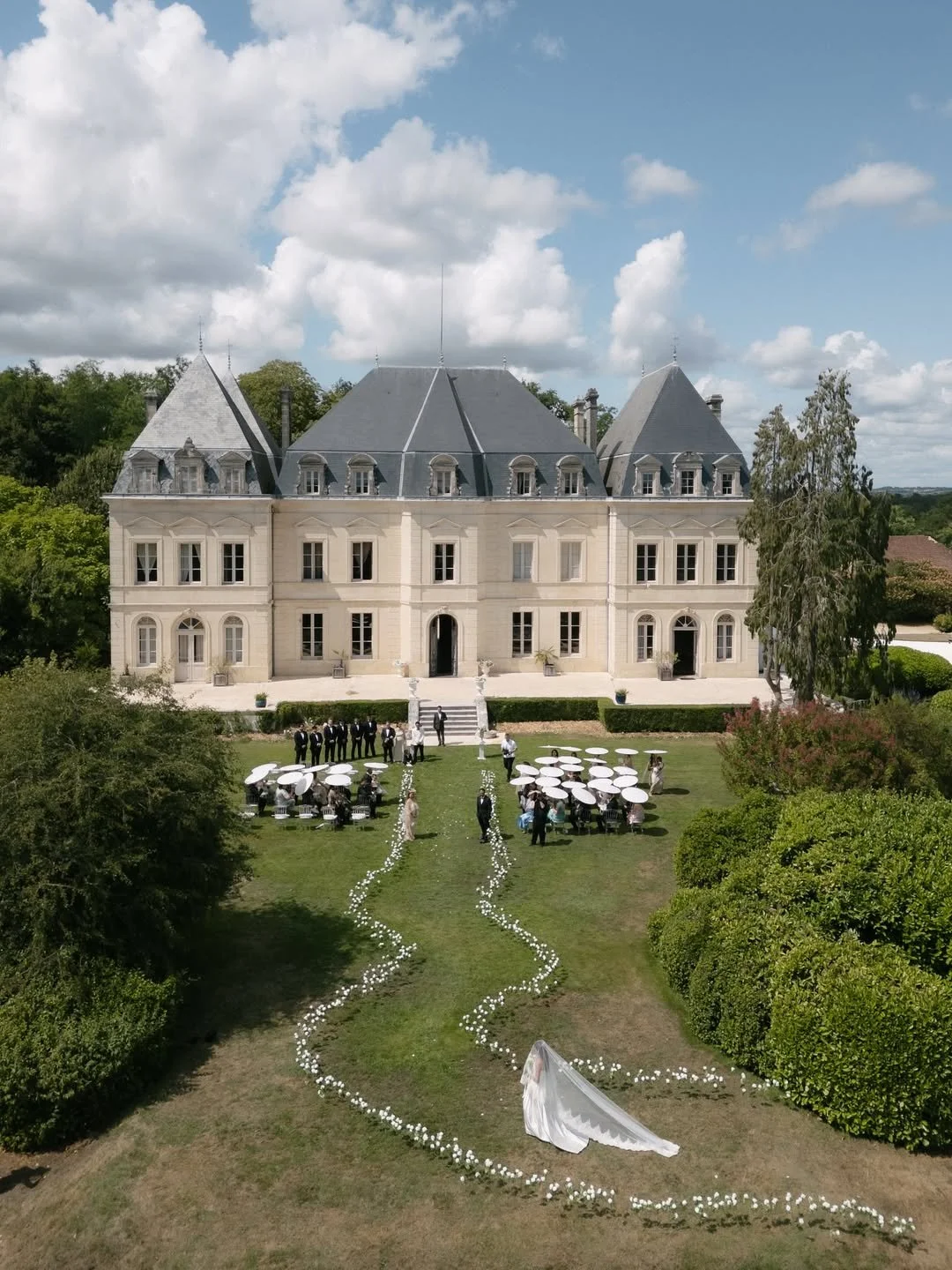 Un mariage se déroule devant un grand manoir avec des invités assis sous des parasols blancs, une mariée en robe blanche marche sur une allée décorée de fleurs, le tout dans un jardin verdoyant avec un ciel partiellement nuageux.