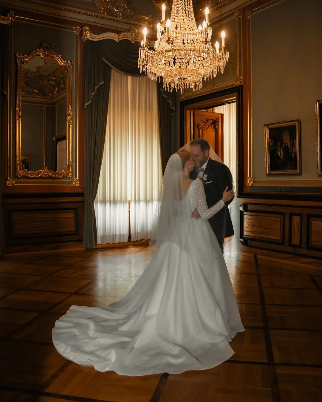 Un couple de mariés qui s'embrassent dans une salle élégante avec un chandelier en cristal suspendu et des rideaux en velours.