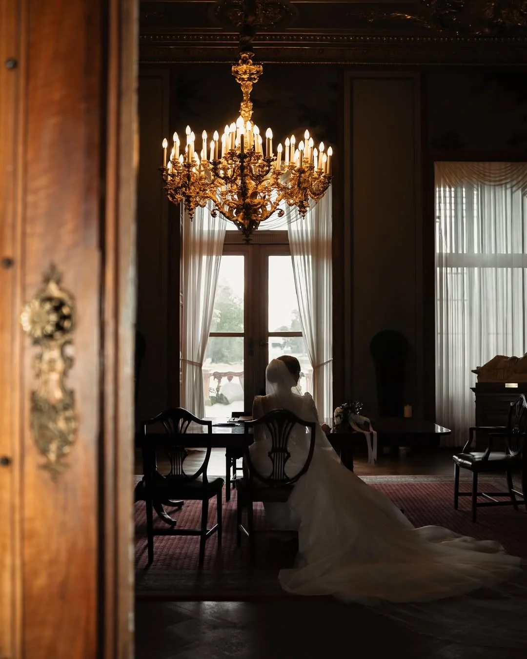 Une mariée en robe blanche assise dans une salle élégante, éclairée par un chandelier doré, avec des rideaux blancs derrière une fenêtre.