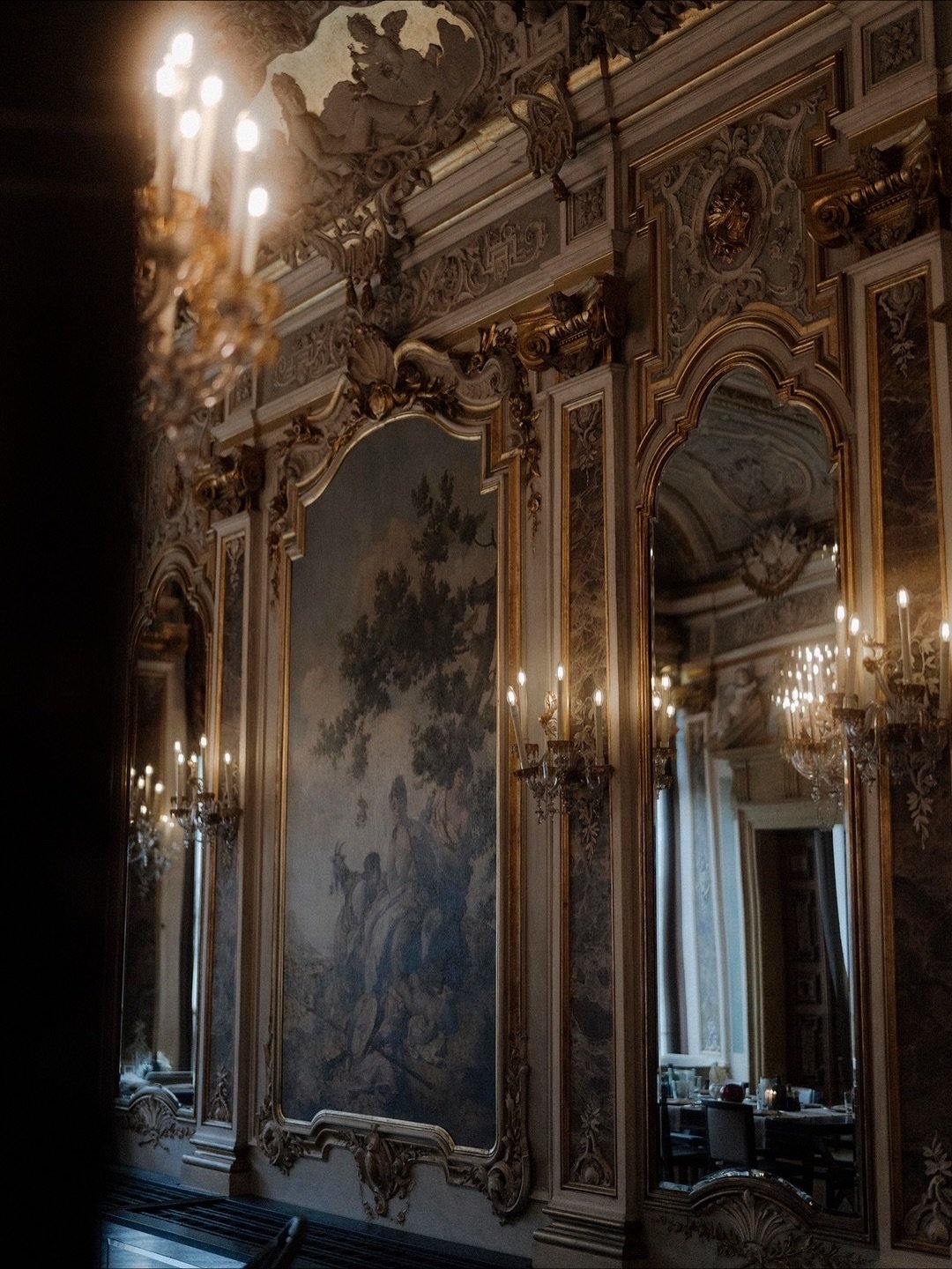 Intérieur de salon richement décoré avec des miroirs ornés, des moulures en or, des chandeliers et un grand tableau peint représentant un arbre et des figures humaines.