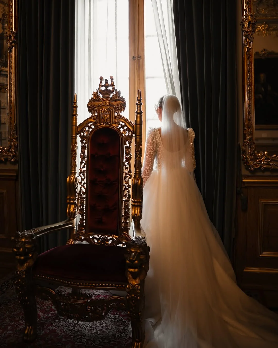 Femme en robe de mariée regardant par une fenêtre dans une pièce luxueuse avec des meubles en bois doré et des rideaux sombres.