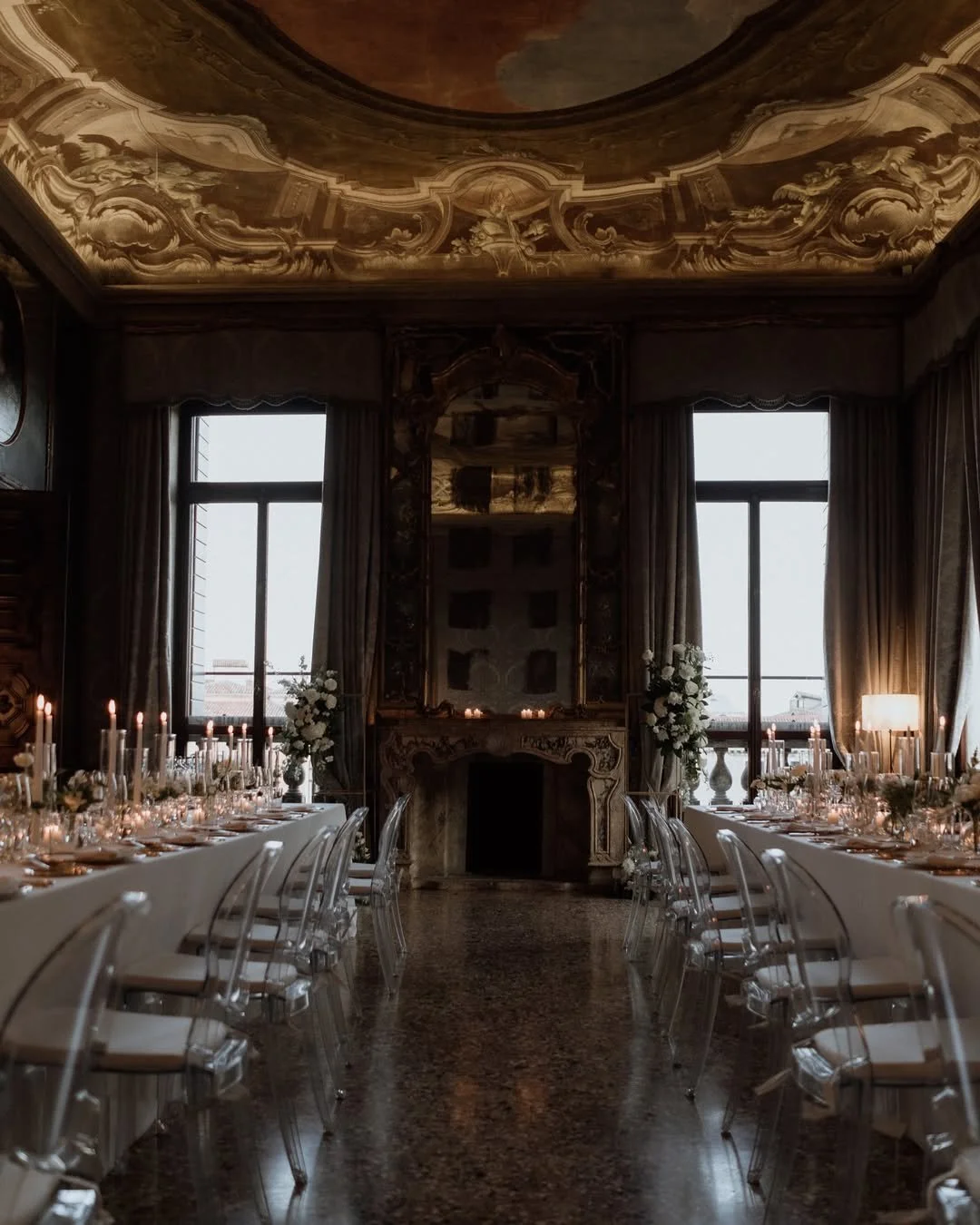 Salle de réception élégante avec deux longues tables décorées de chandeliers et de fleurs blanches, grand miroir ancien au-dessus de la cheminée en marbre, fenêtres avec rideaux en velours, plafond peint avec des fresques baroques.
