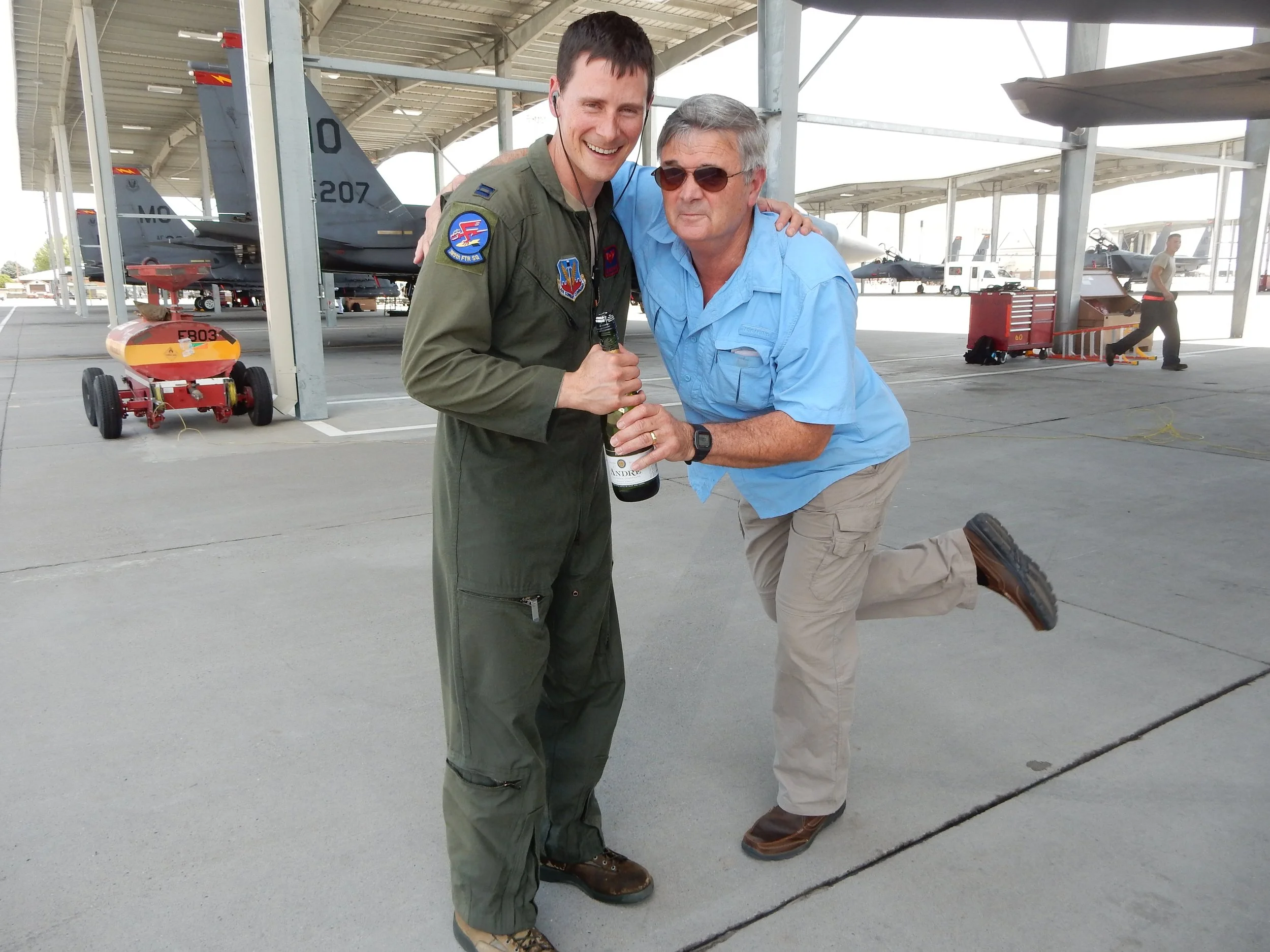 Two men, one in military pilot uniform and the other in casual clothes, are celebrating with a bottle of champagne at an airfield with fighter jets in the background. One man is holding the champagne bottle, and both are smiling and posing for the photo.