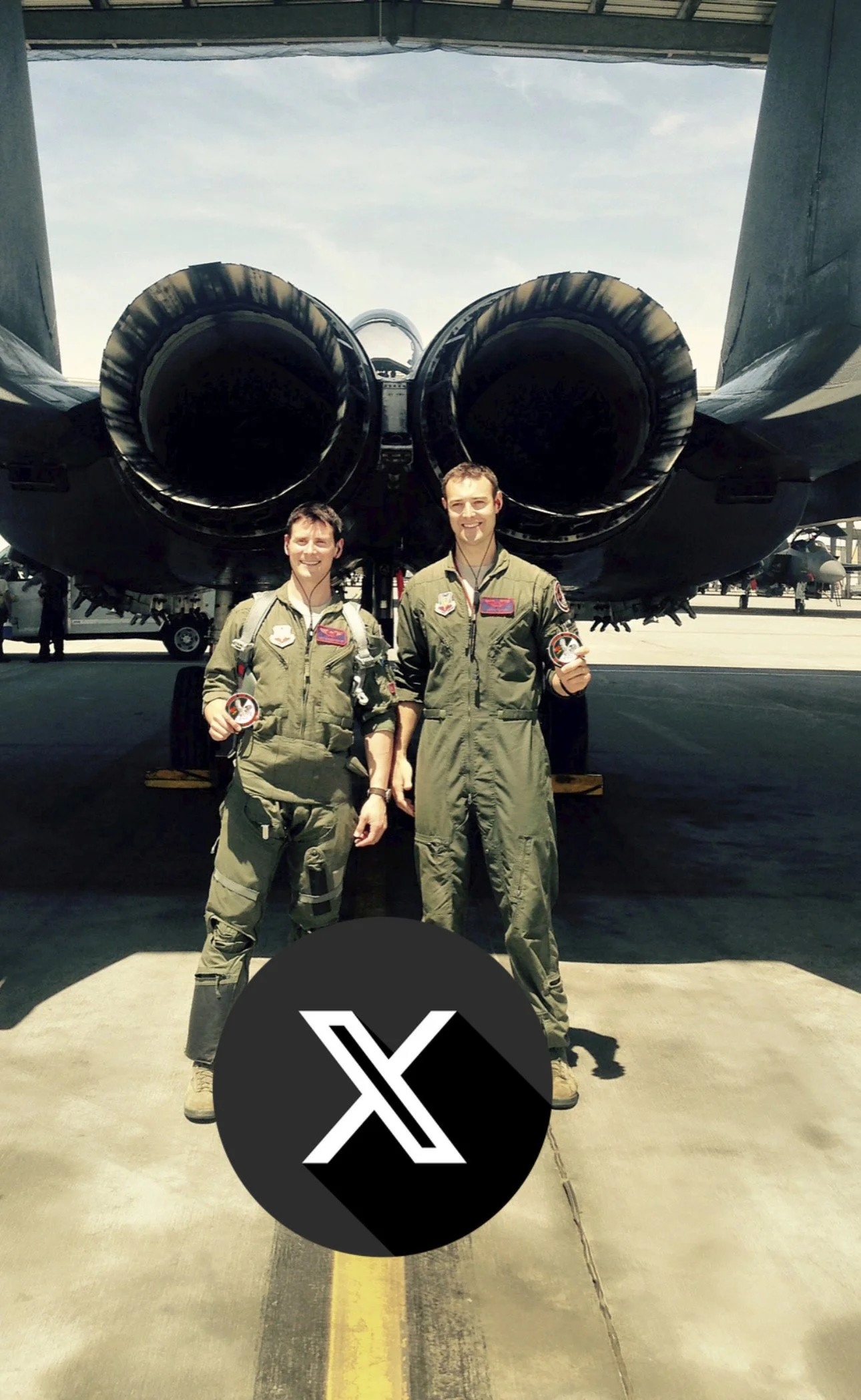 Two pilots in flight suits standing in front of a large military aircraft with two jet engines, underneath an aircraft hangar roof.