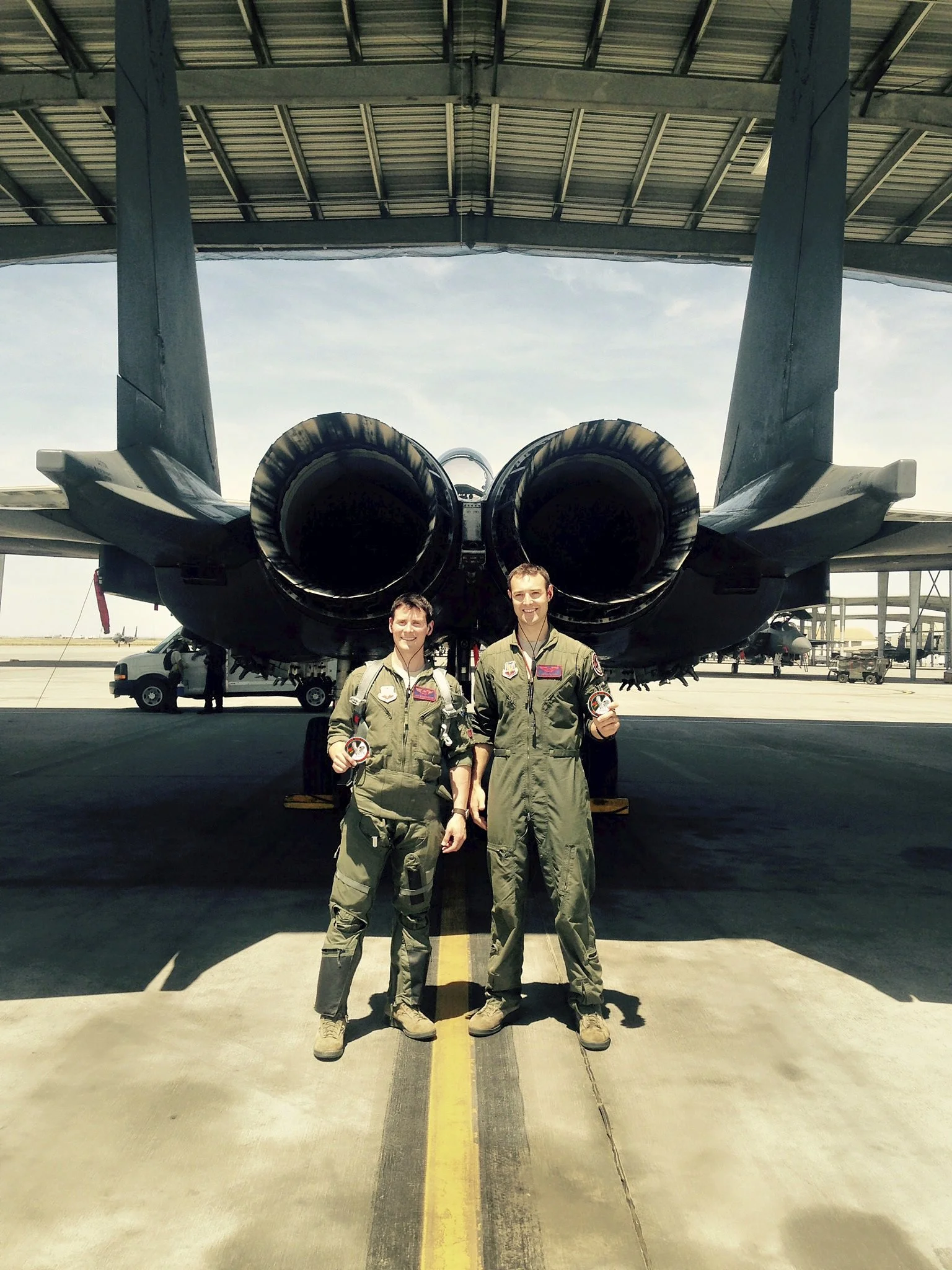 Two pilots in flight suits standing in front of a jet aircraft inside a hangar.