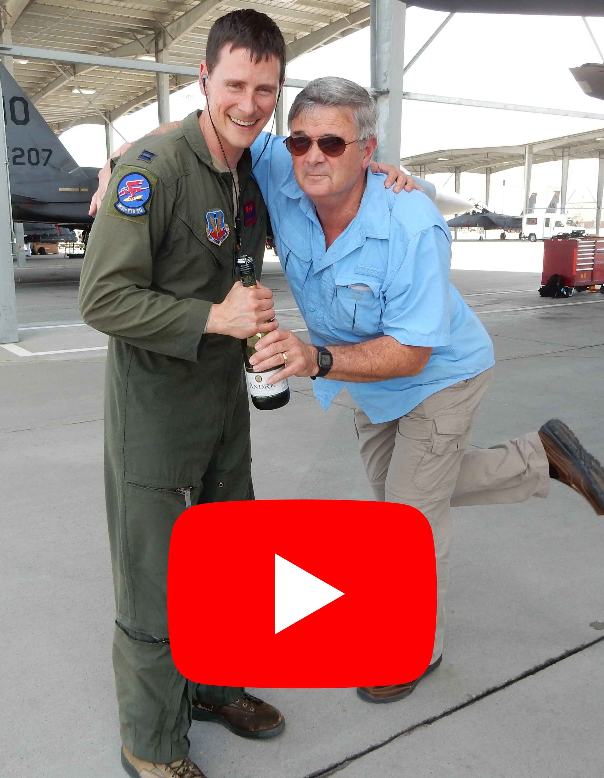 Two men, one in military flight suit and one in casual attire, pose together at an airbase with aircraft in the background. They are smiling, and the man in casual clothes is holding a bottle of wine.