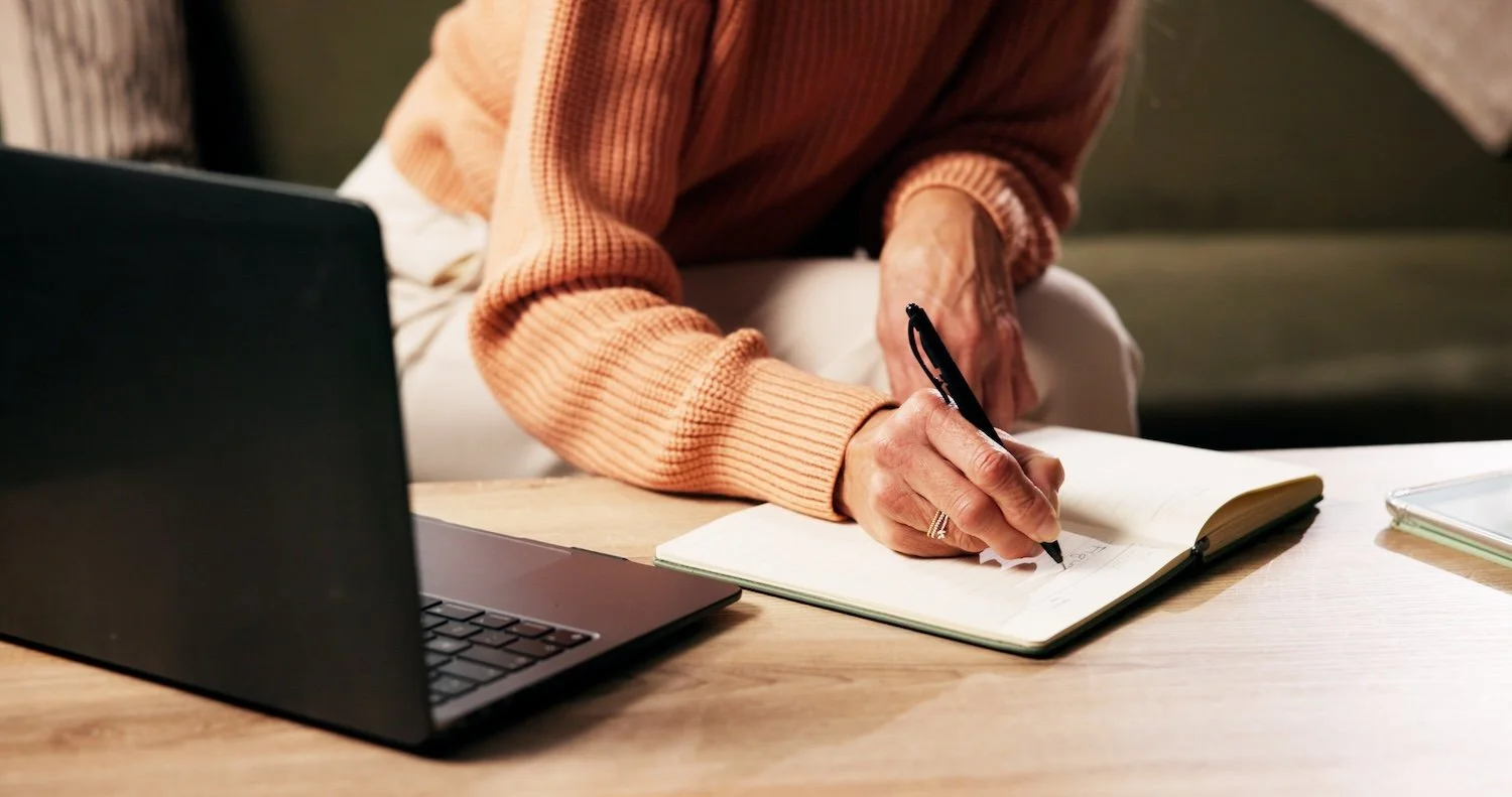 A person wearing a peach sweater writing in a notebook on a desk, with a laptop and a tablet nearby.
