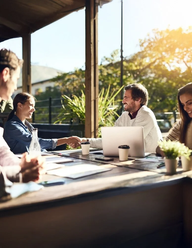 Business meeting with five people at a wooden table outdoors, two people shaking hands, laptops, coffee cups, and a sunny background with trees.