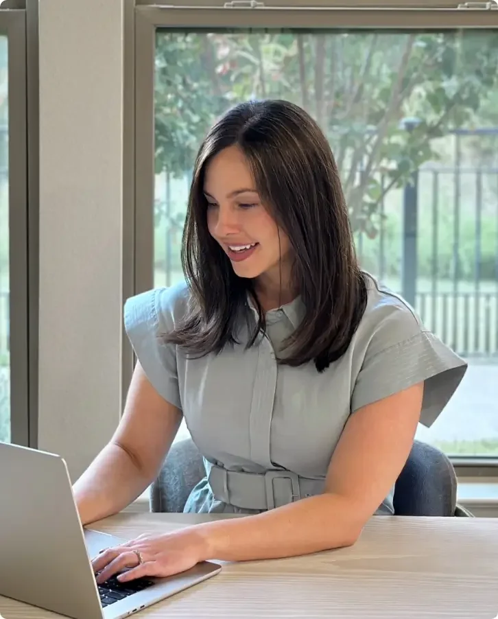 Samantha Scott working on a laptop at a table in a room with large windows and greenery outside.