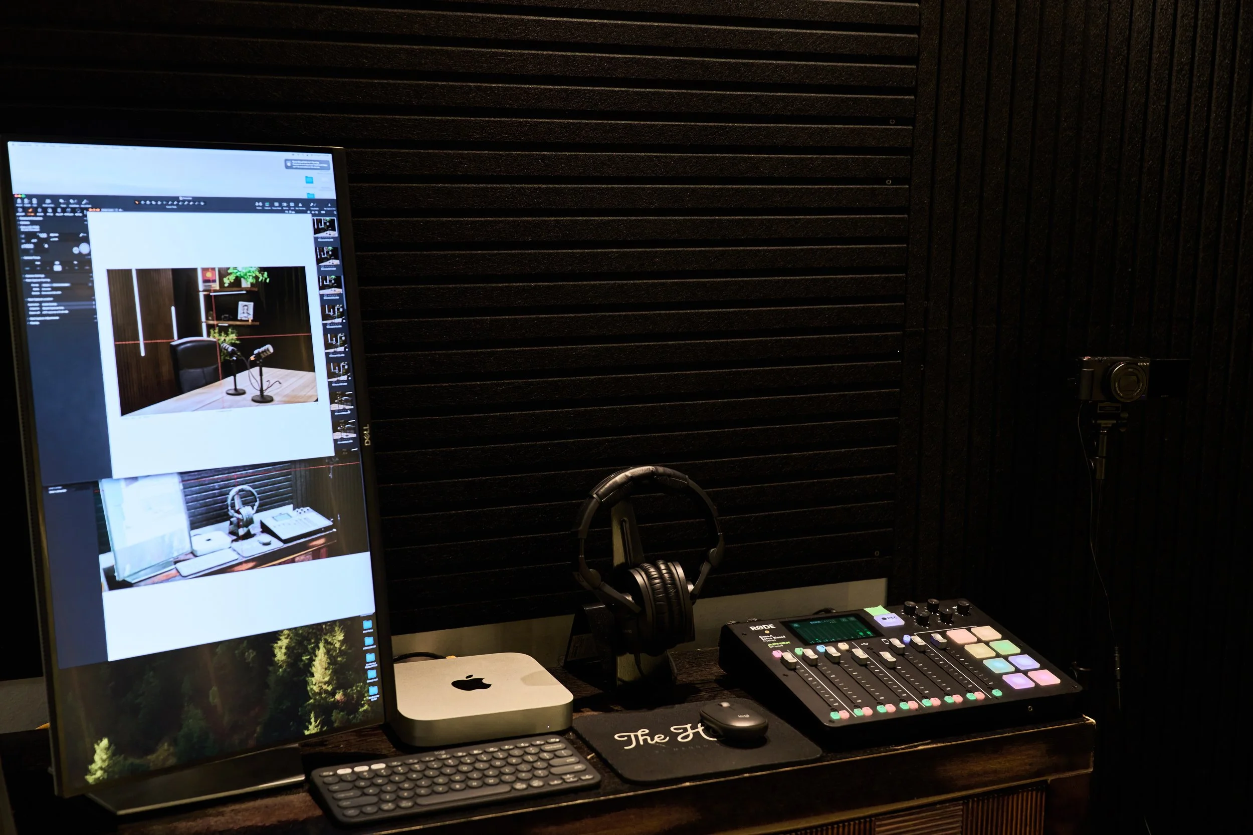 A recording or podcasting station with a computer monitor, headphones, a small audio mixer, software, a keyboard, a mouse, and a mini desktop computer on a wooden desk, with black soundproofing foam on the wall behind.