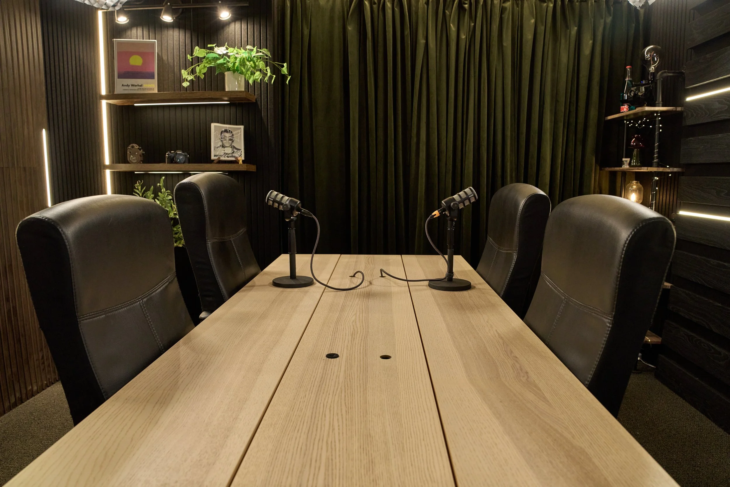 A modern podcast recording studio with a wooden table, four black leather chairs, two microphones on stands facing each other, a dark curtain backdrop, and wall shelves holding decor items like a potted plant, framed pictures, and bottles.
