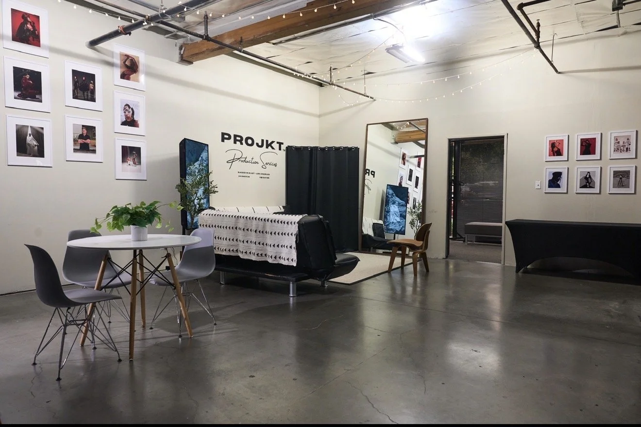 An indoor photography studio with a seating area, featuring framed photos on the walls, a table with a plant, a large mirror, and photographic equipment, including a backdrop with a privacy curtain, and string lights hanging from the ceiling.