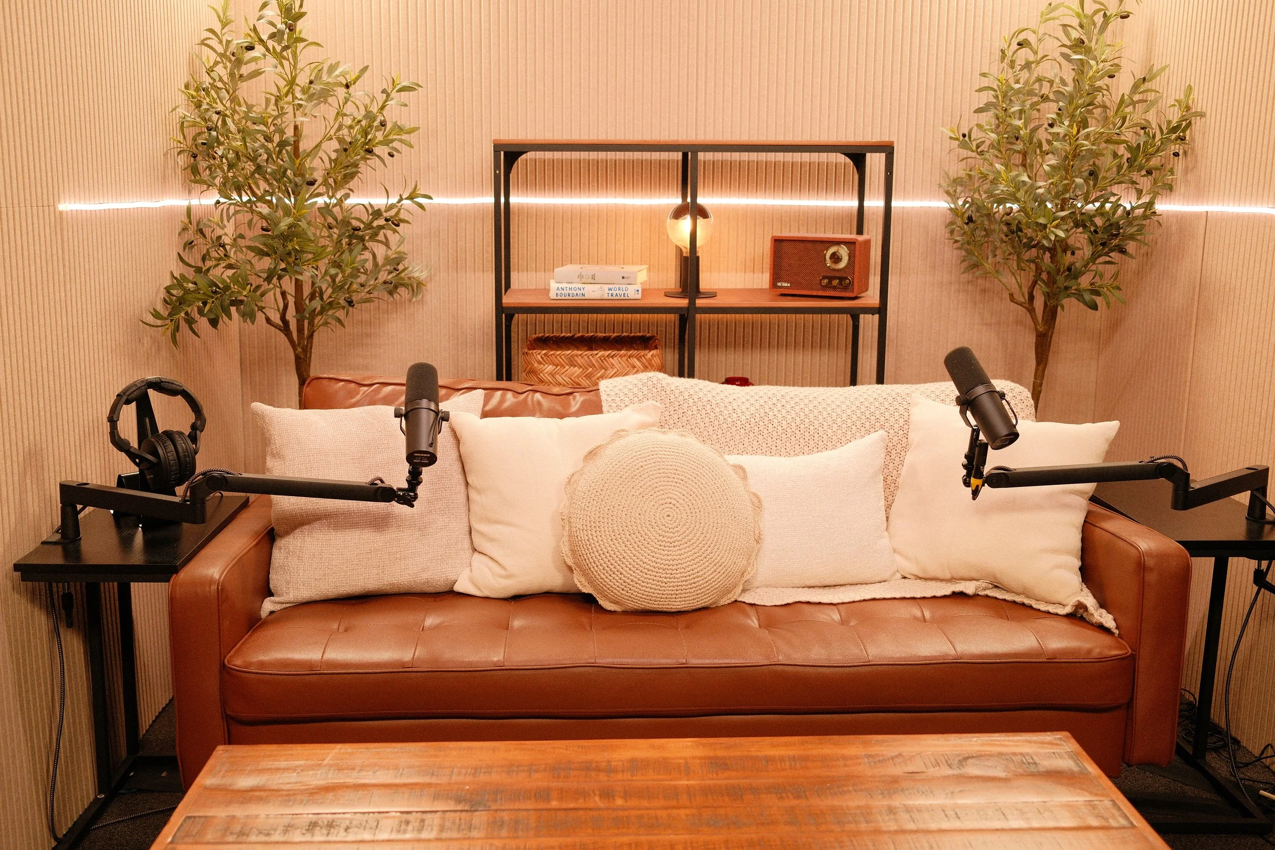 A cozy podcast recording setup with a brown leather sofa, multiple cream and textured pillows, two microphones on adjustable stands, a side table with headphones, a wooden table, two potted trees, a black shelving unit with books, a lamp, a vintage radio, and a decorative woven basket, all against a warm beige ribbed wall.
