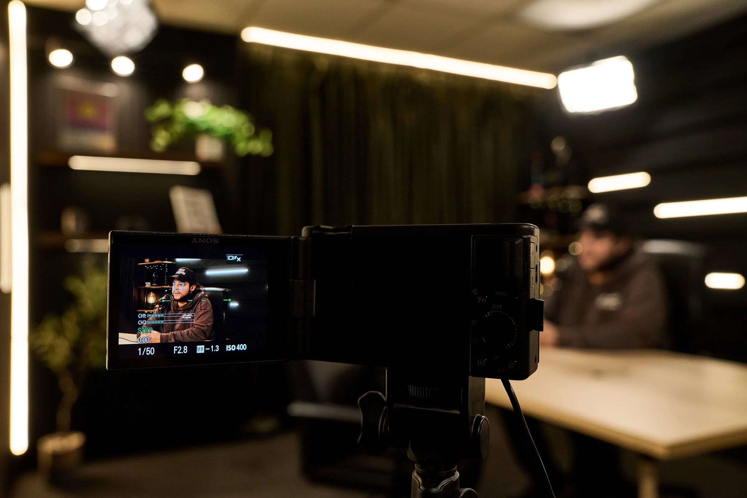 Video camera capturing an interview with a person wearing a cap and dark clothing in a dimly lit room with modern lighting and decor.